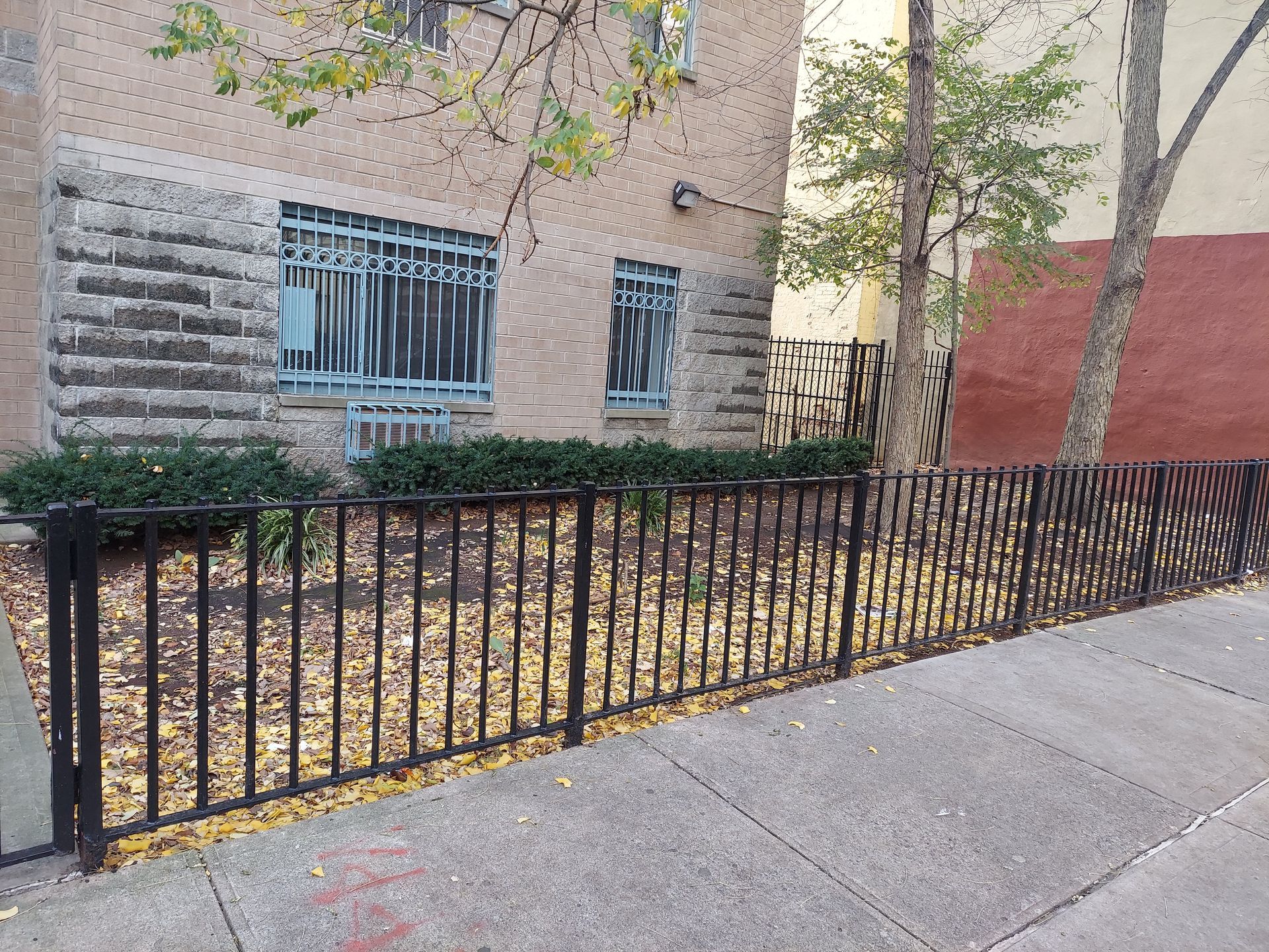 Brick building with barred windows, black metal fence, and sidewalk lined with trees and shrubs.