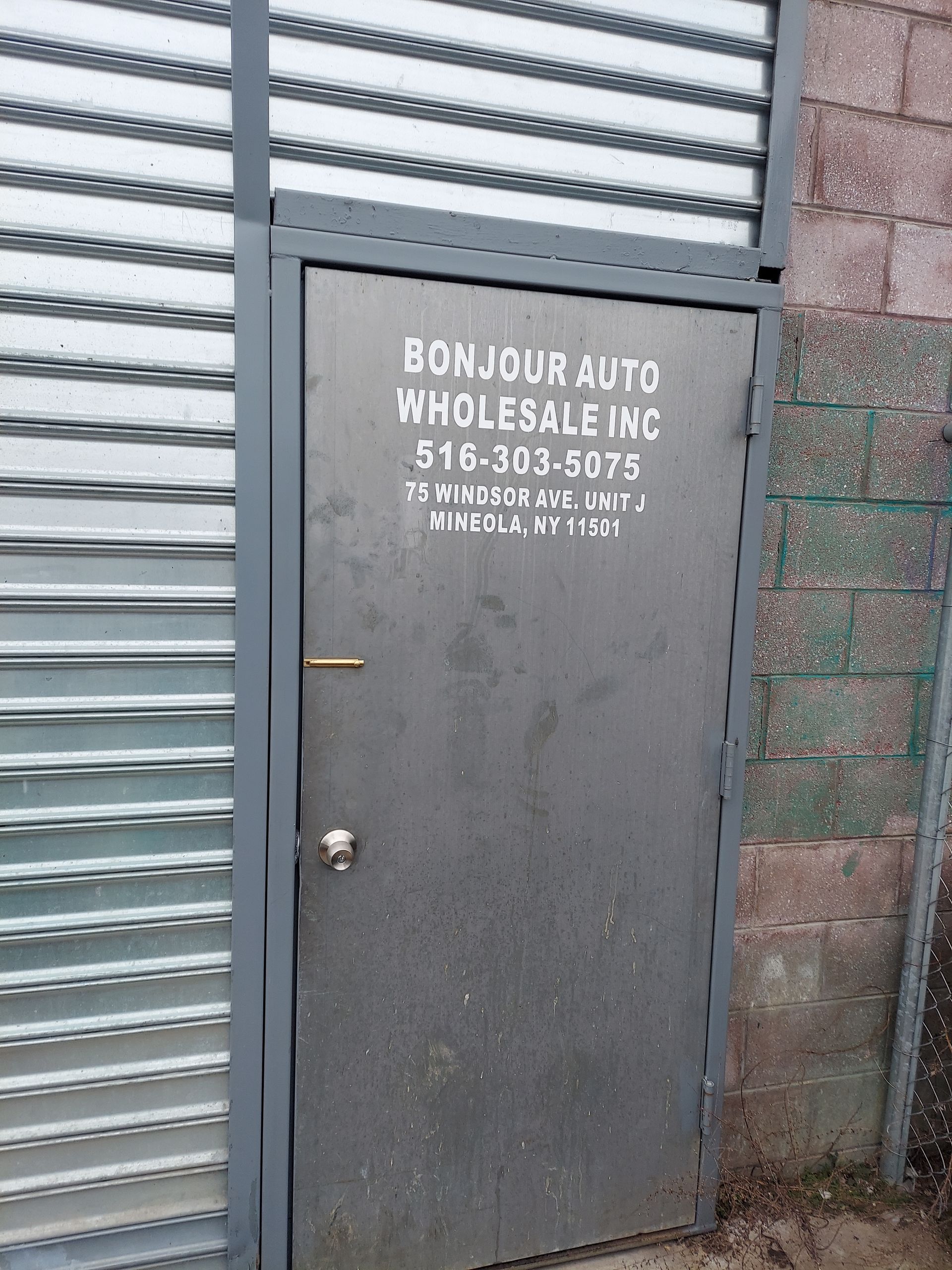 Weathered gray metal door with “BCM Auto Wholesale LLC” painted on it beside a corrugated wall.