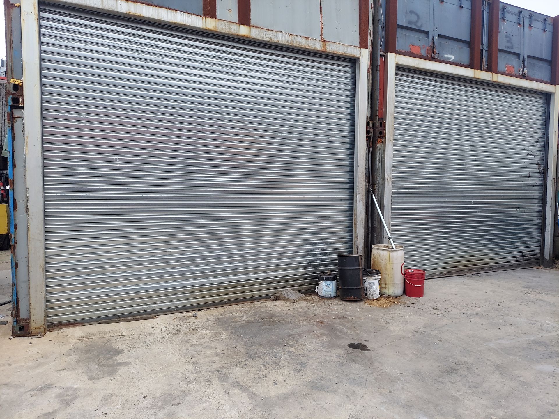 Two corrugated metal garage doors on a dusty street, with buckets and a red can by the right door
