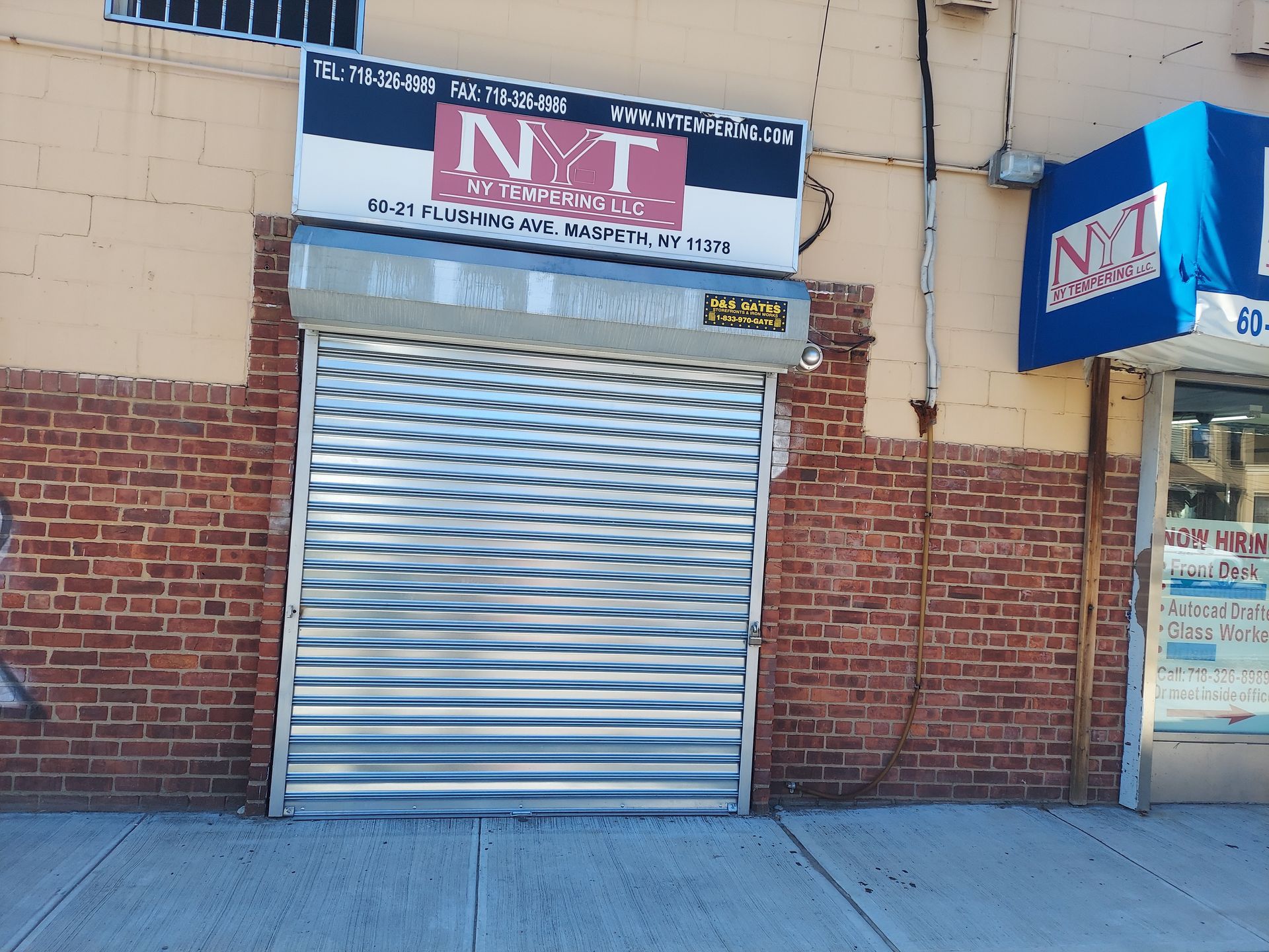 Closed storefront with a silver roll-up door and red-and-white signage above, beside a brick wall.