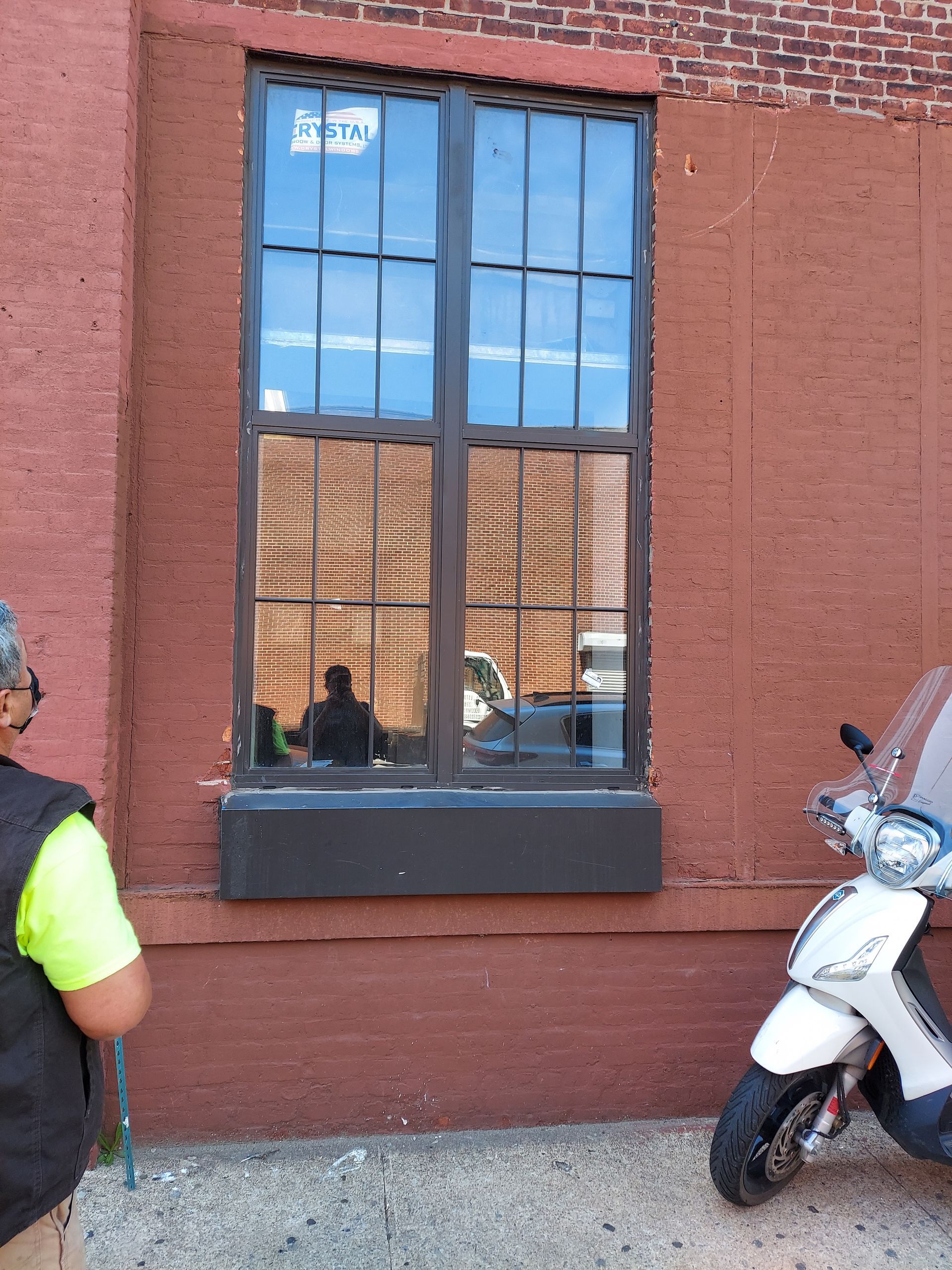 Brick building facade with tall window reflecting a parked scooter and street scene