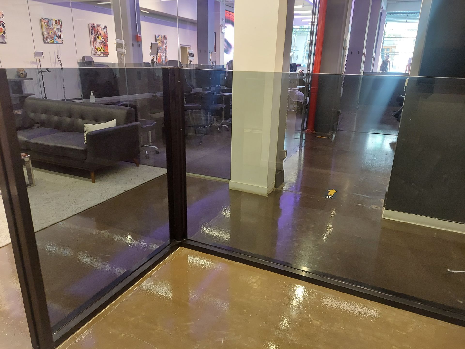 Glass-walled office hallway with a couch visible inside and polished floors reflecting light.
