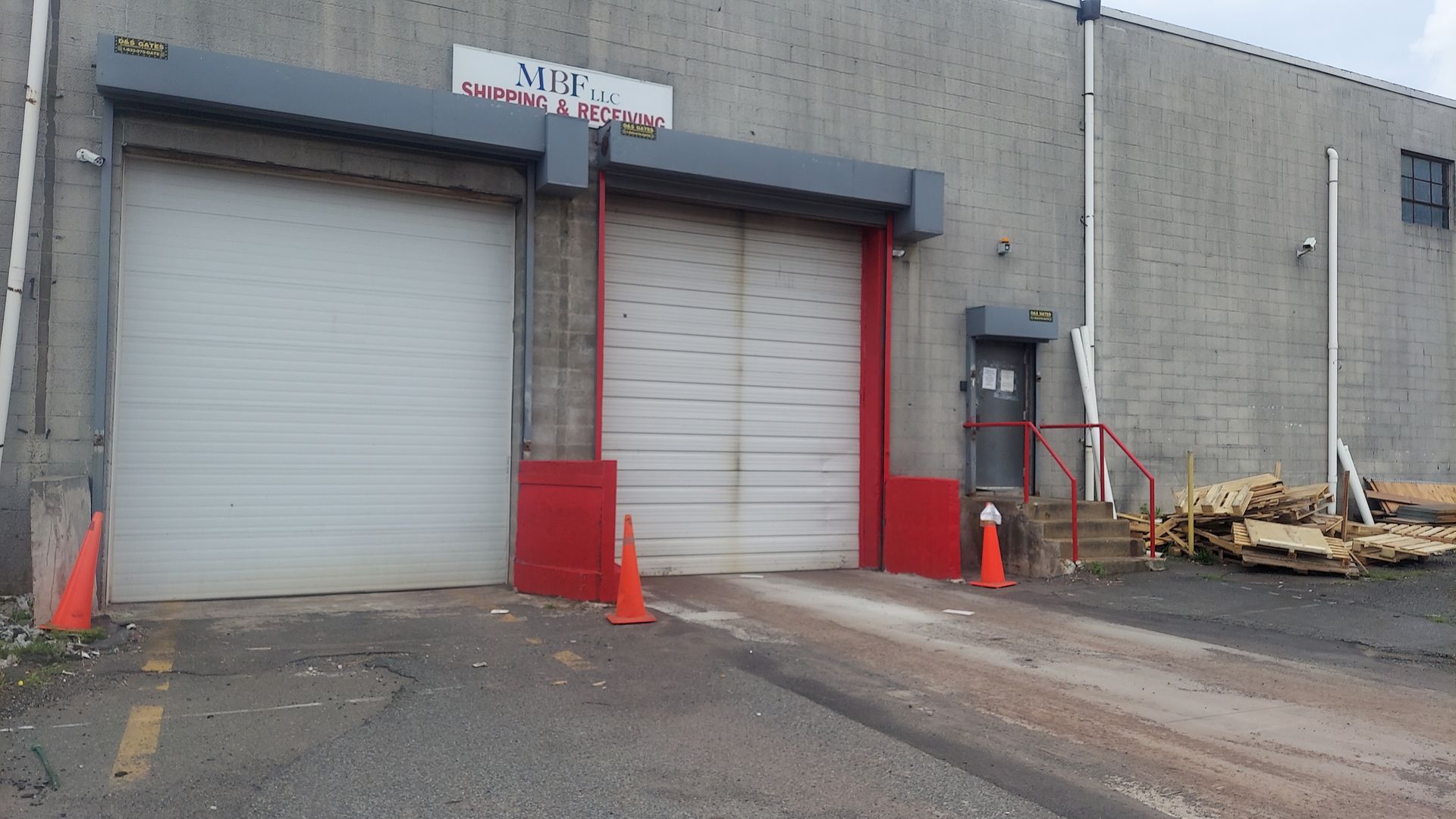 Industrial loading dock with two roll-up doors, red safety cones, and a small stairway beside a gray building.