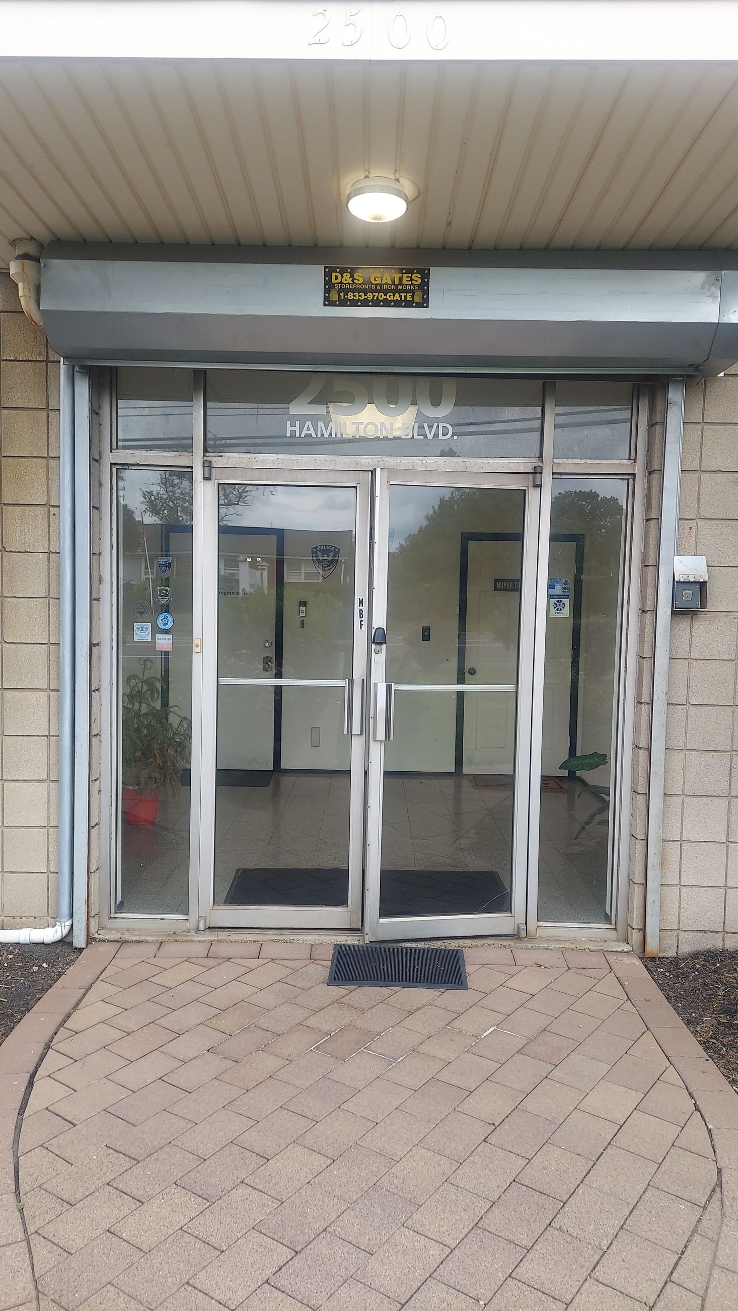 Glass entrance doors under a covered entryway at a building, with brick paving and a wall-mounted intercom.