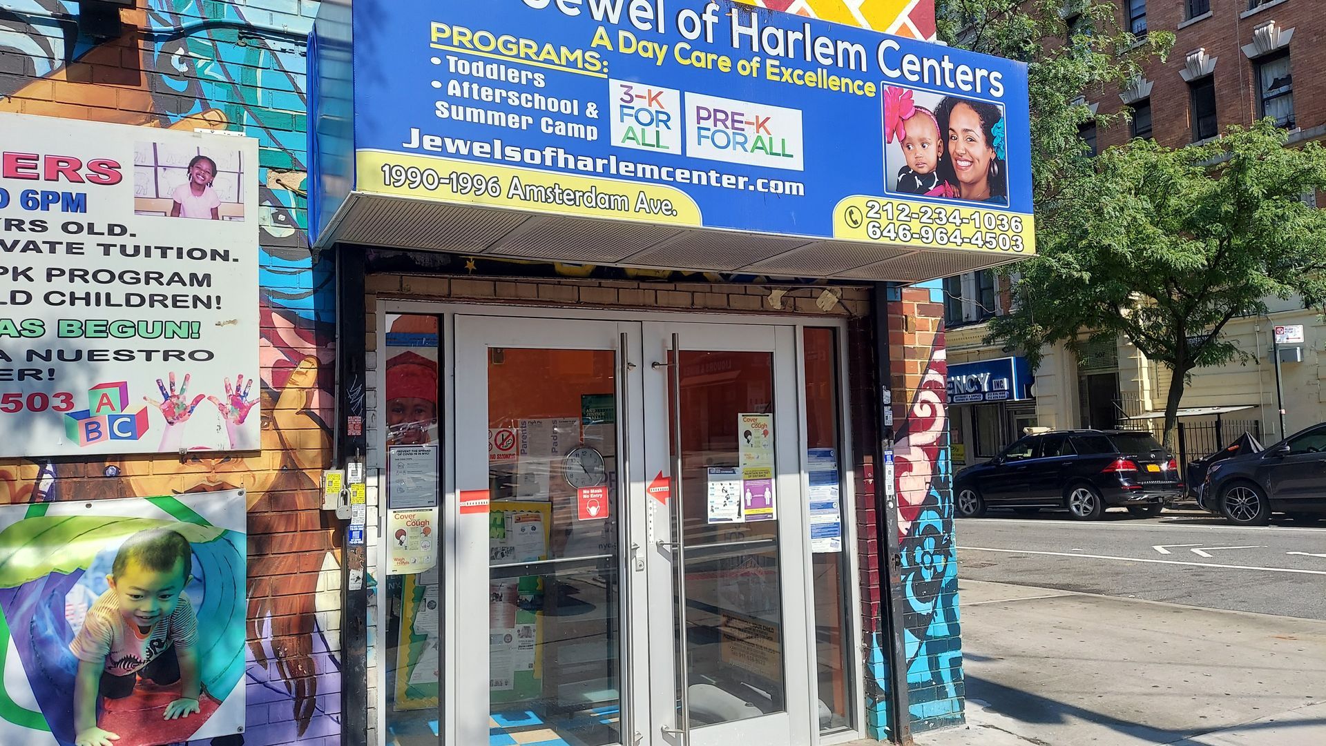 Storefront with a blue sign, glass double doors, and colorful posters on a sunny city sidewalk.
