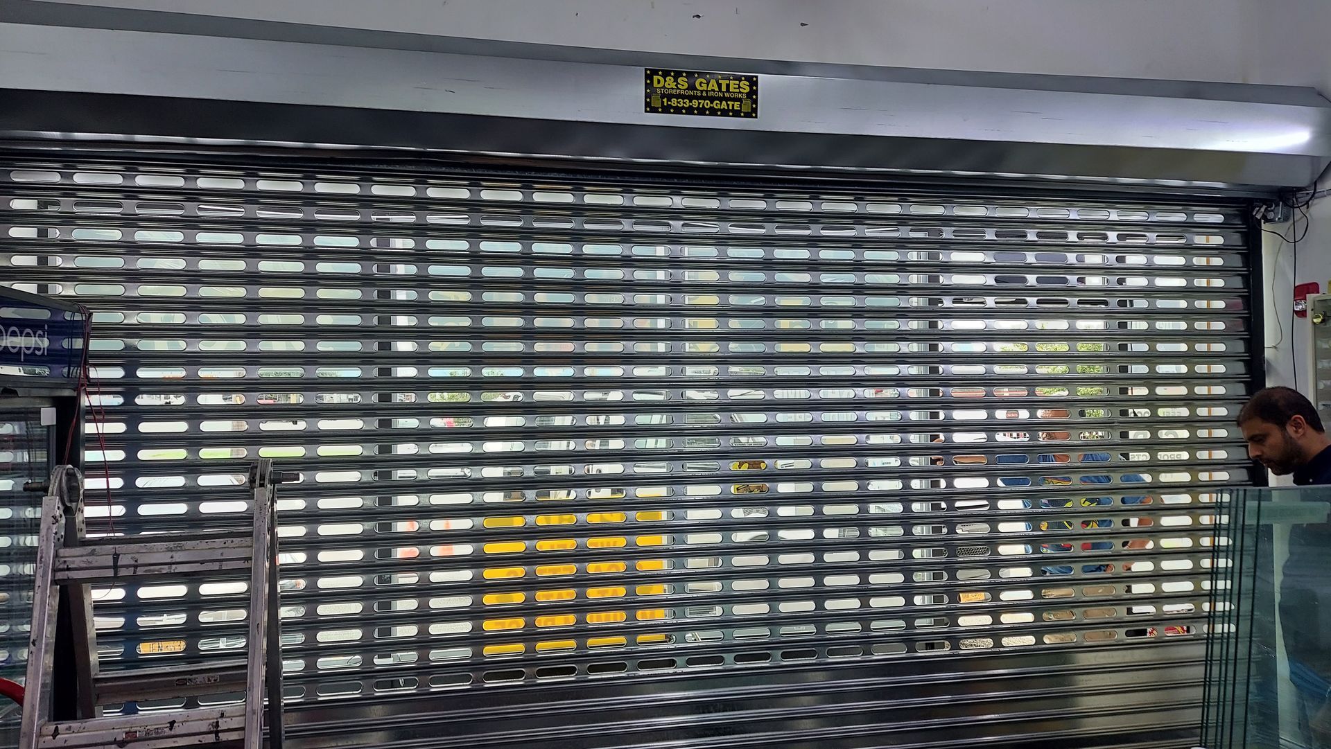 Closed metal shutter across a storefront, with a man standing at the right edge near the entrance.