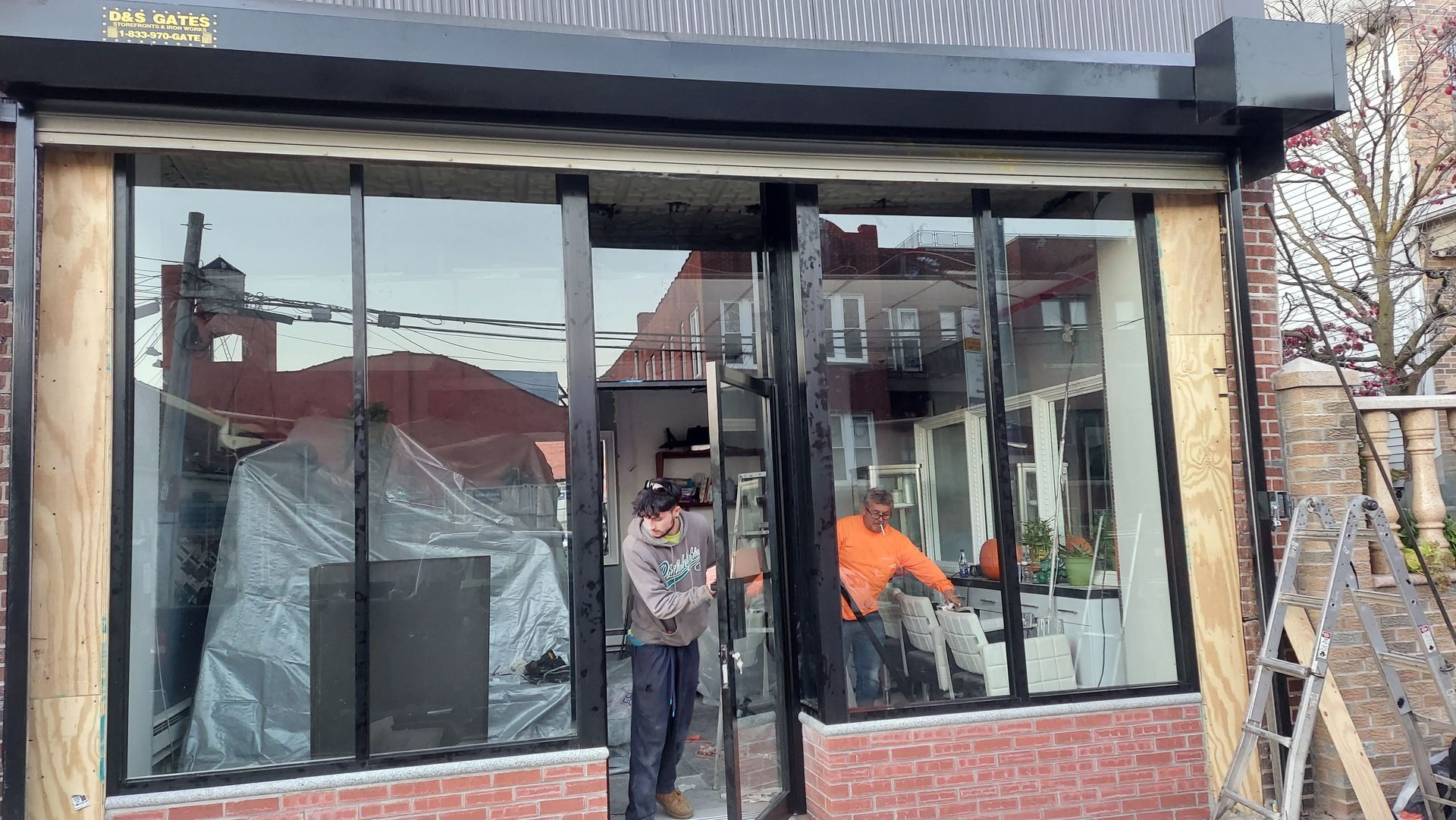Two workers install large glass panels on the front of a storefront under construction.