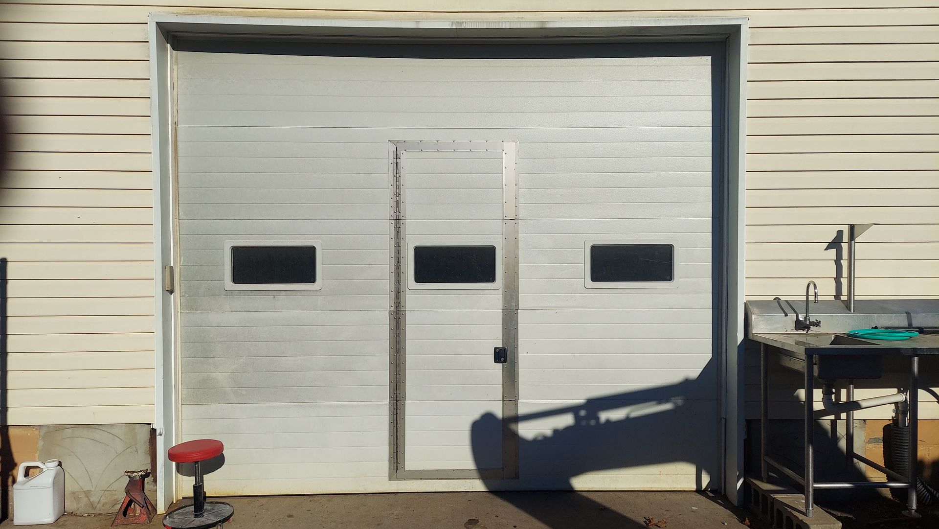 White garage door on beige building with three narrow windows and a small door, with yard equipment in front.