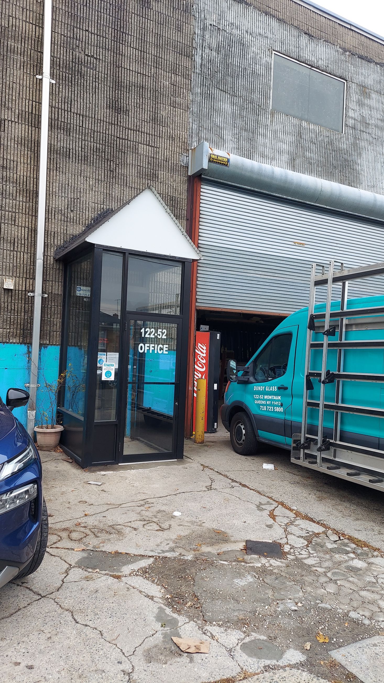 Small glass entrance beside a blue truck and parked car on a gravel lot by a corrugated wall
