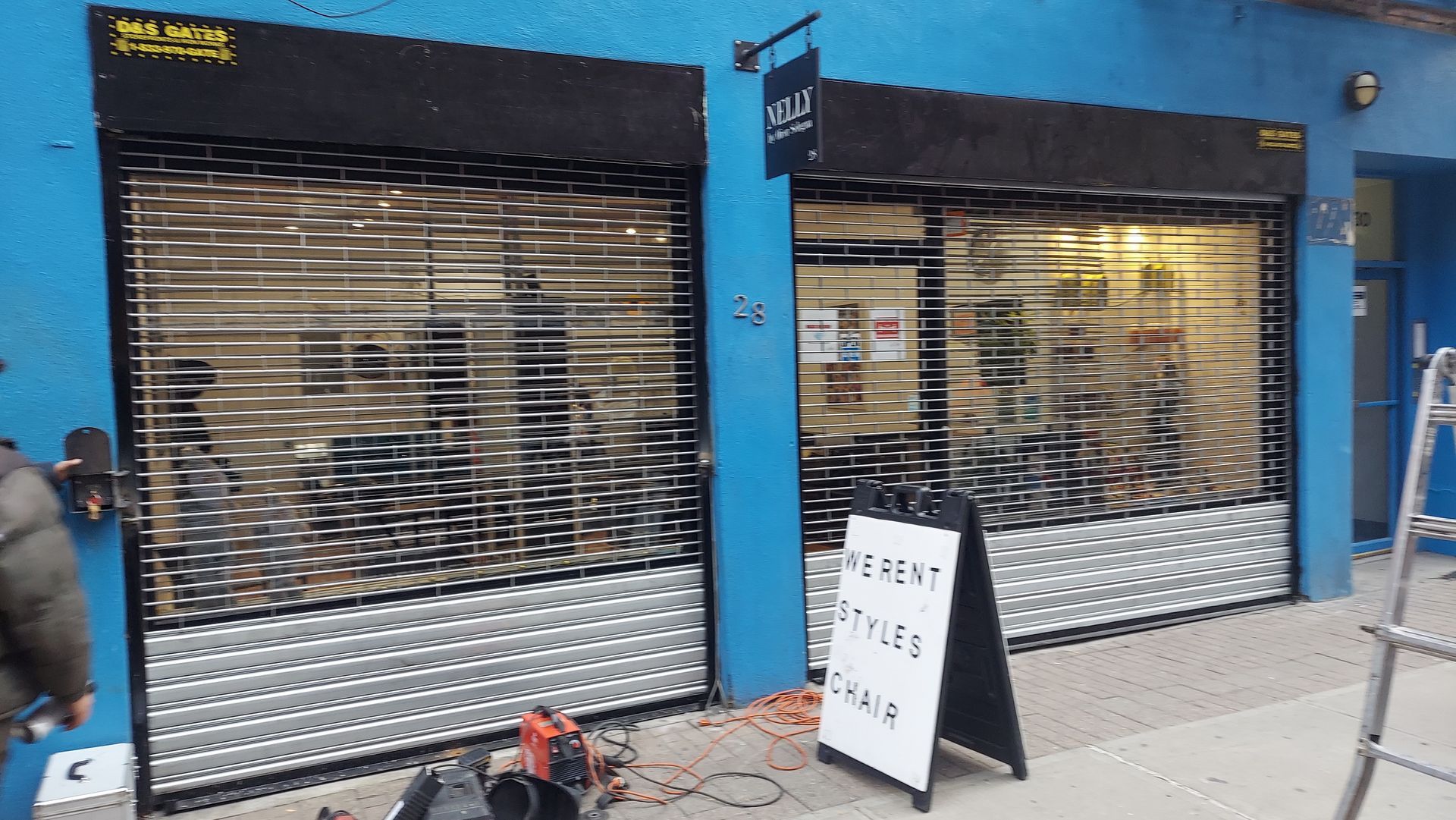 Closed storefronts with silver shutters on a blue building, and a sidewalk sign outside