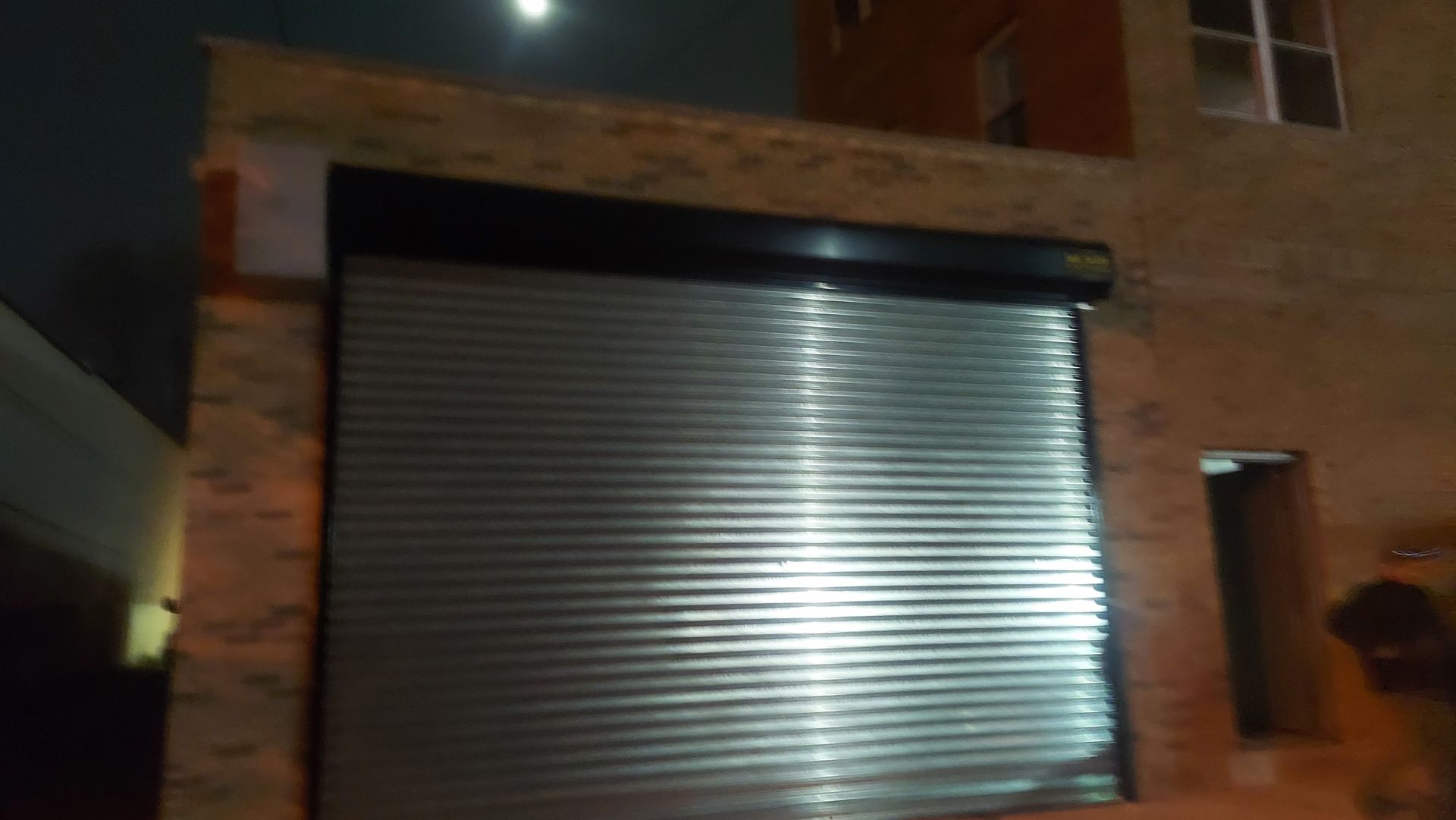 Closed metal garage door on a brick building at night, lit by a bright light.