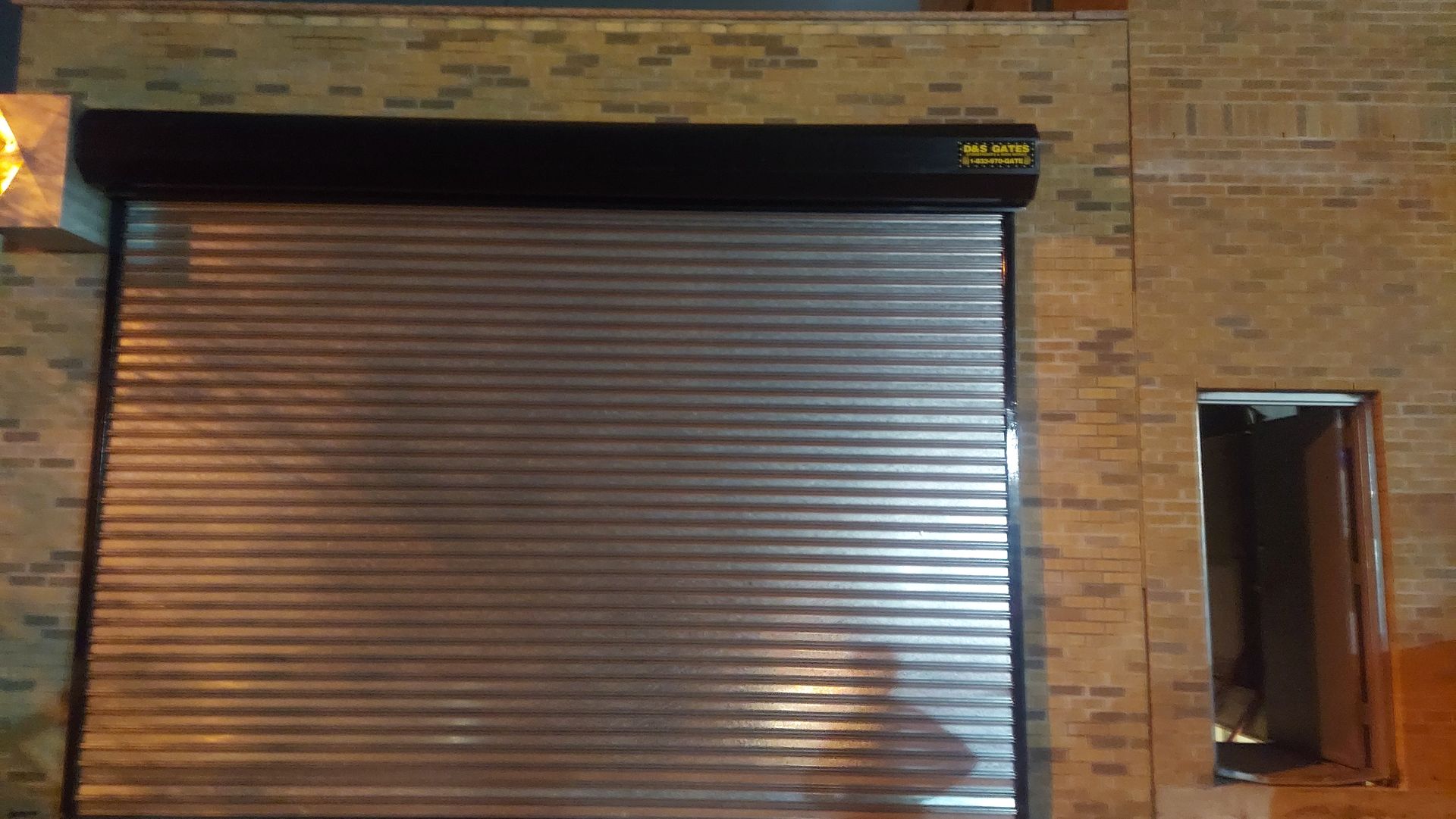 Closed metal roll-up door in a brick wall, with a small open doorway to the right.