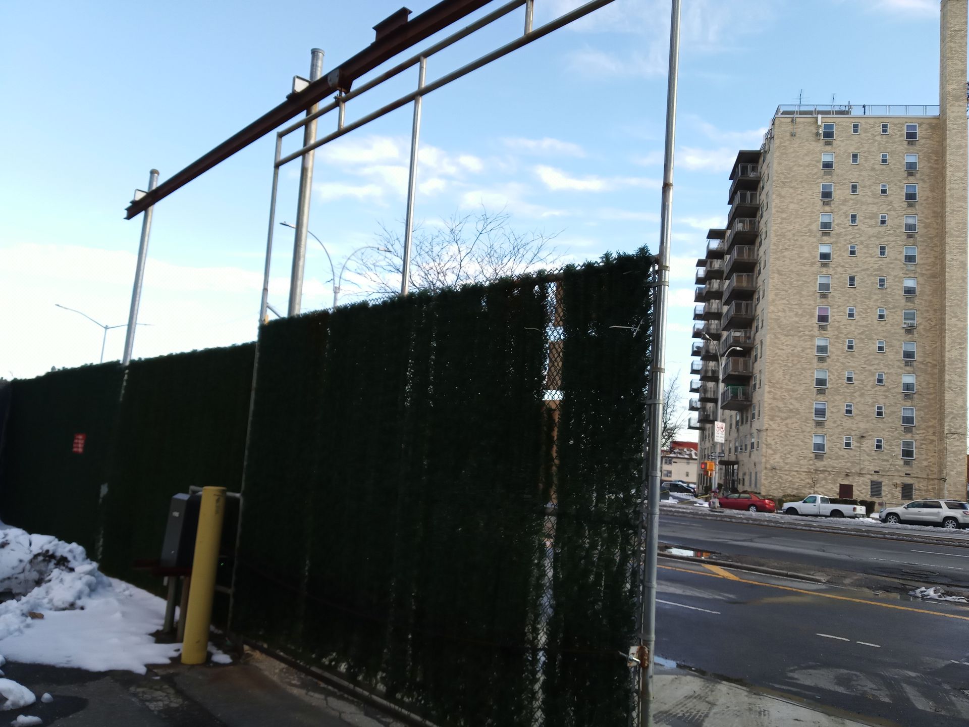 Snowy urban street with tall apartment building and green fence under a clear blue sky