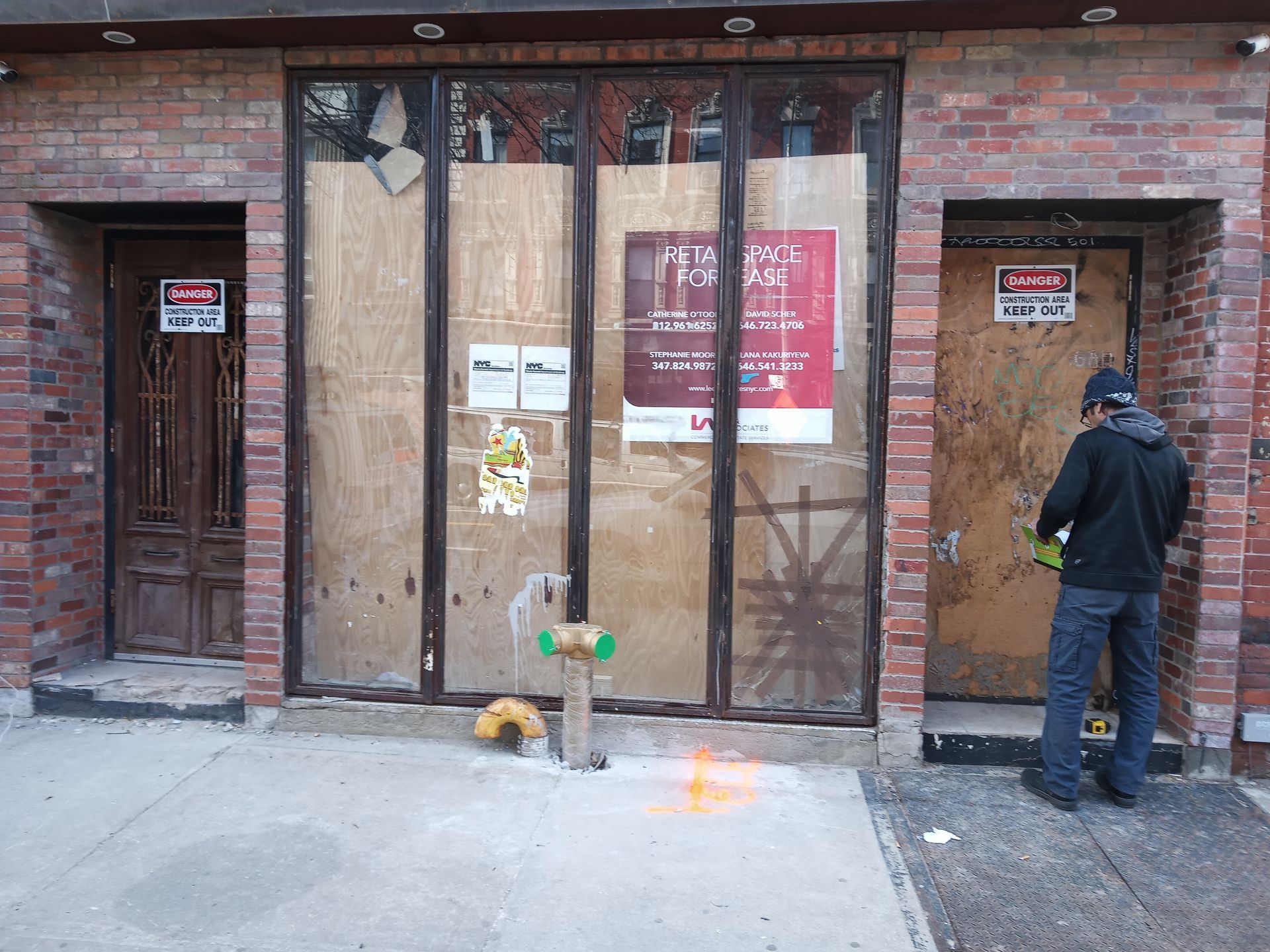 A boarded-up storefront with glass doors, warning signs, and a man standing on the sidewalk checking his phone.