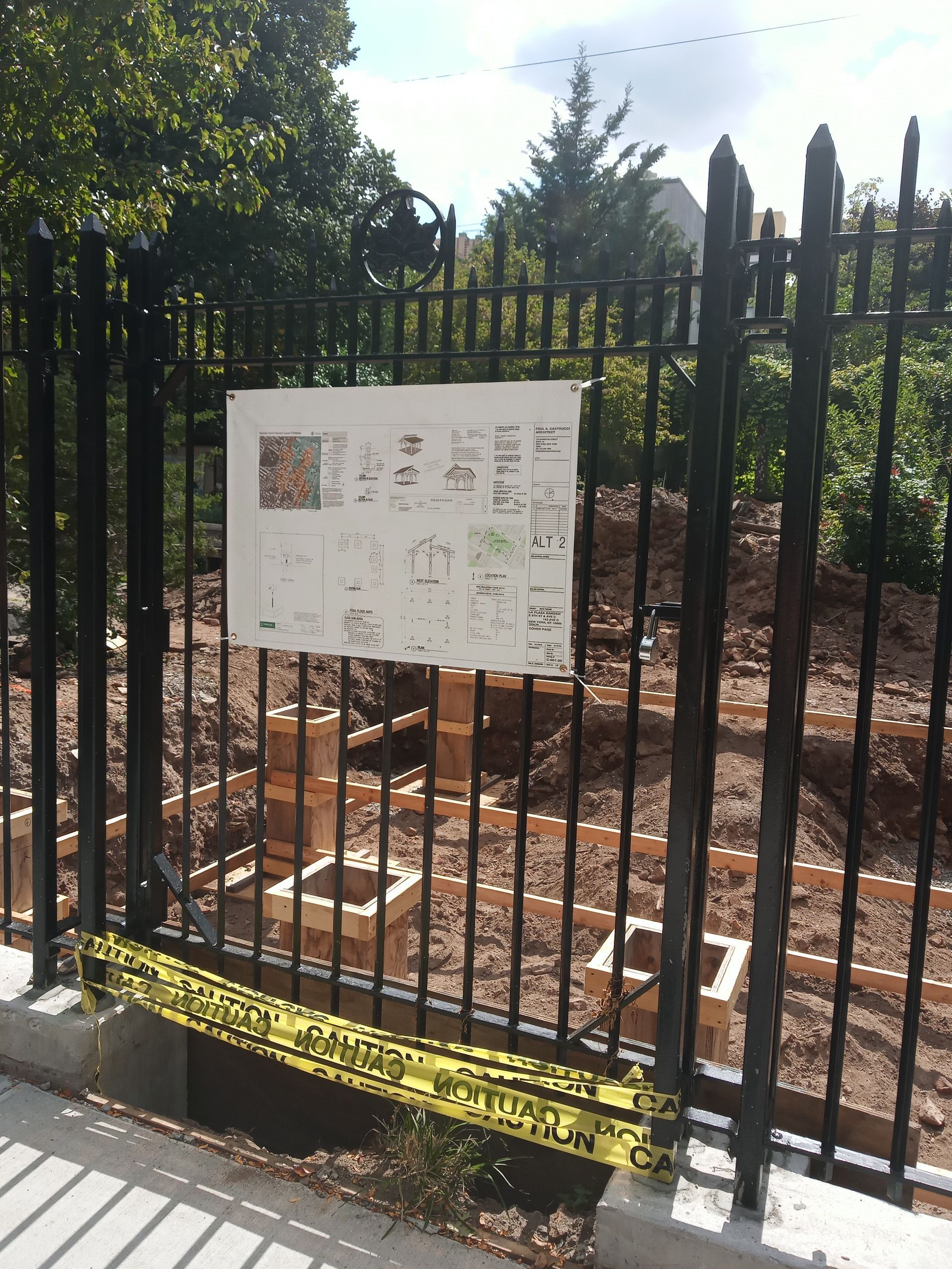 Black metal fence with a posted notice in front of a brick-covered lot and trees beyond.