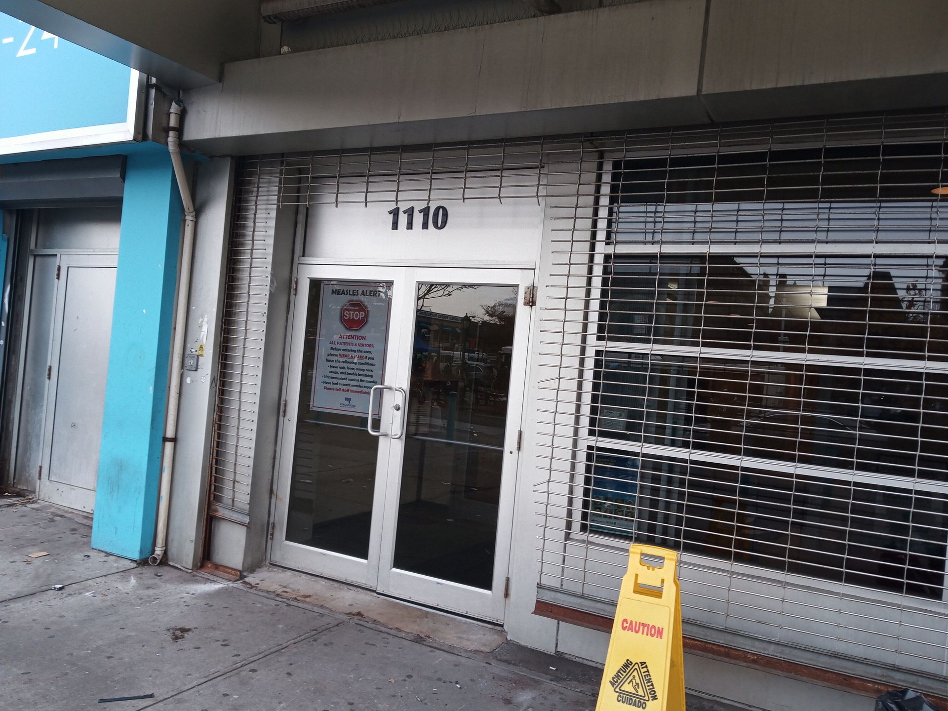 Storefront entrance with glass doors, metal security gate, and a yellow caution sign on the sidewalk
