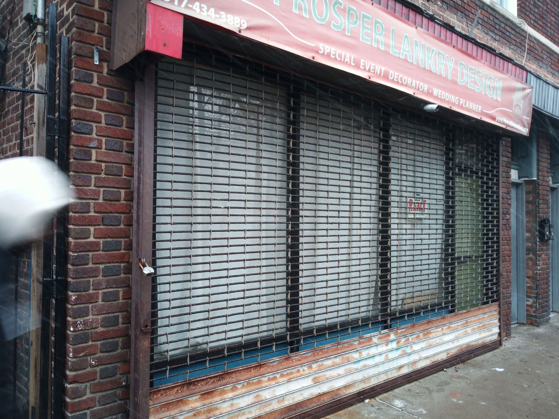 Closed metal storefront shutters with faded red awning on a wet street.