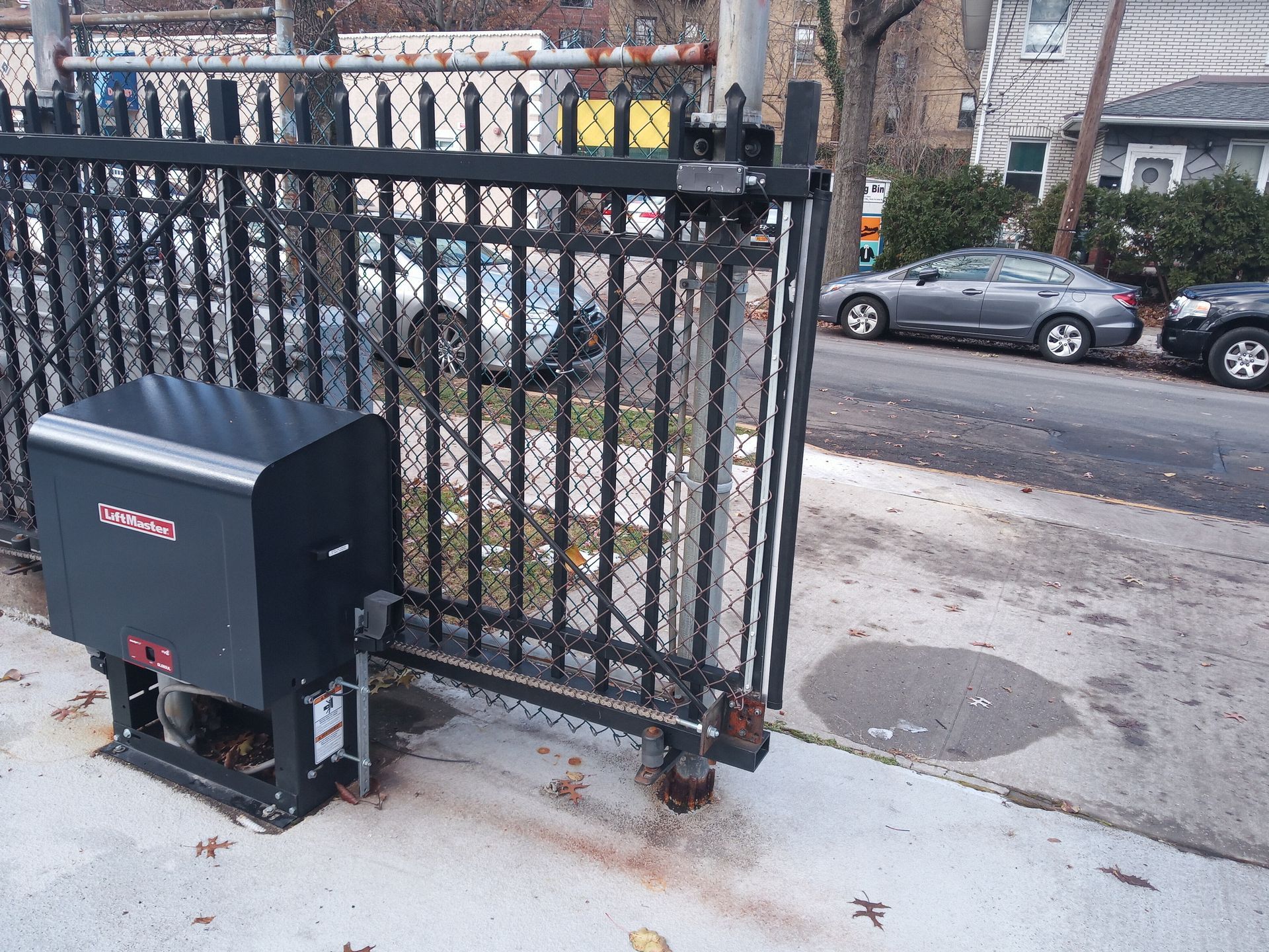 Black metal fence beside a sidewalk, with a black utility box and parked cars across the street.