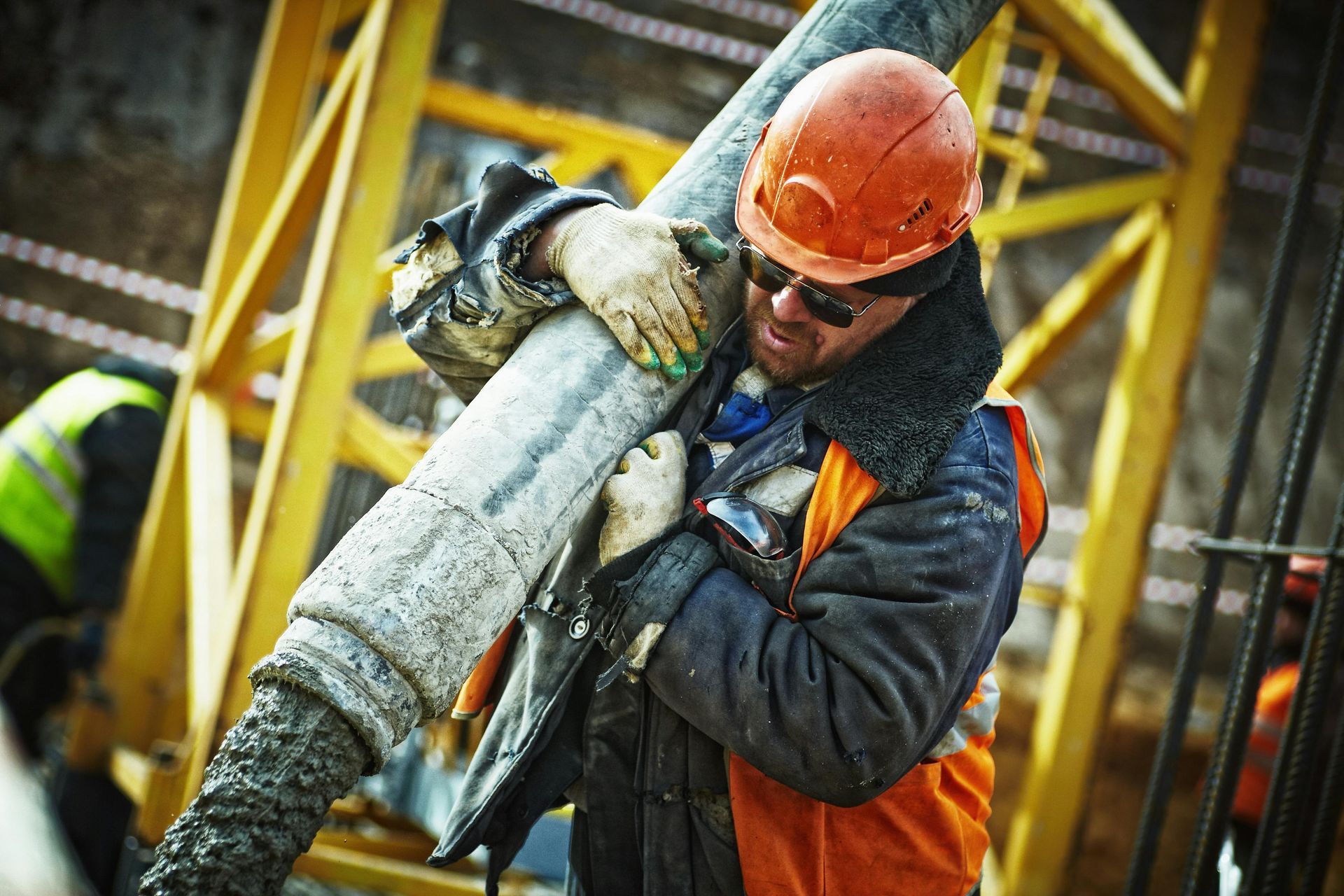 Worker in orange hard hat and vest carrying a heavy pipe at an industrial site.