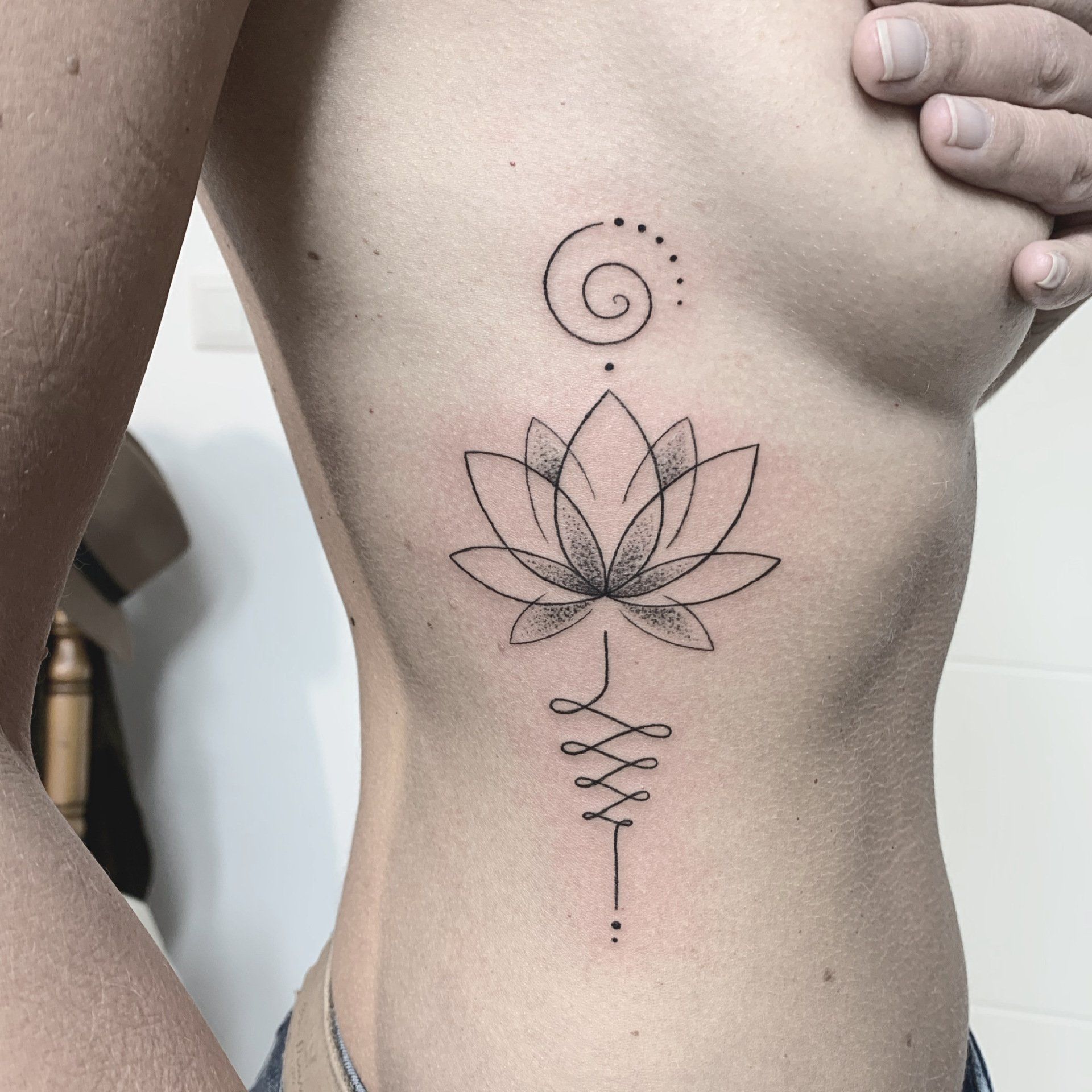 Tätowierung einer Lotusblume mit einem geschwungenen Muster darüber und einer stilisierten Schrift darunter an der Seite einer Person.