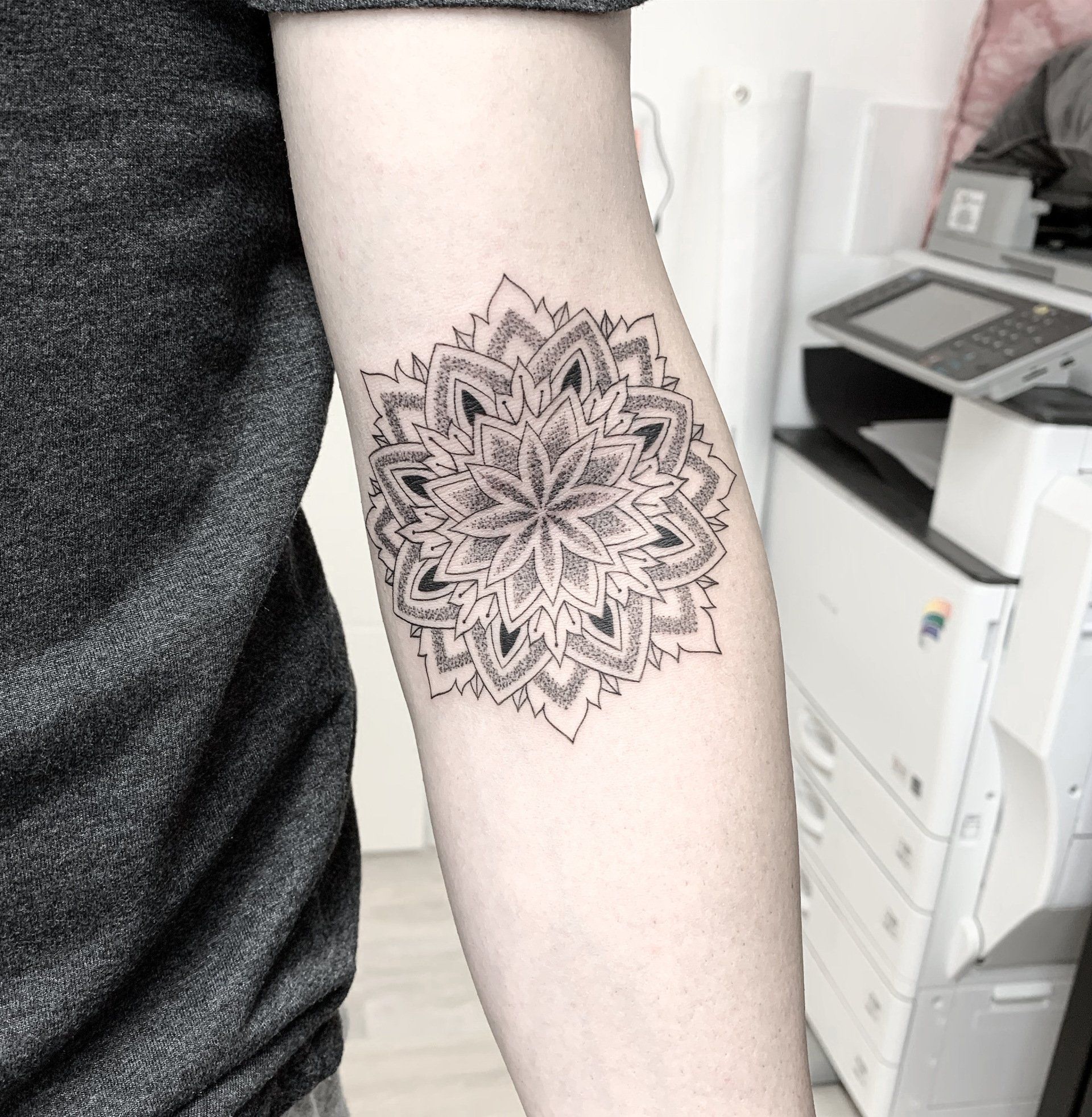 Ein Arm mit einem schwarzen Mandala-Tattoo hebt sich vor einem verschwommenen Hintergrund ab, im Hintergrund ist eine Büromaschine zu sehen.