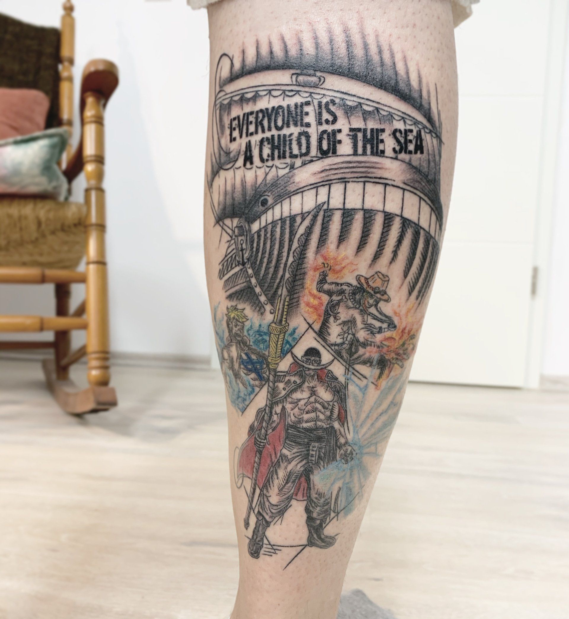 Tätowierung am Bein mit einem Schiff und einer Figur. Text: „Jeder ist ein Kind des Meeres.“ Details im Aquarellstil.