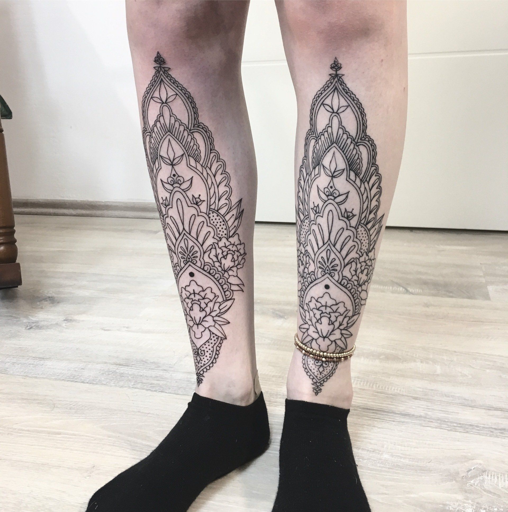 Zwei Beine mit passenden, kunstvollen schwarzen Tätowierungen, die schwarze Socken tragen, stehen in einem Innenraum.