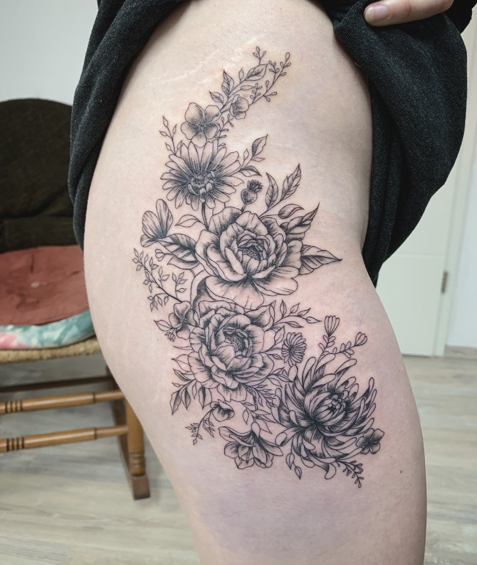 Schwarzes florales Tattoo auf dem Oberschenkel einer Person, mit Rosen und anderen Blumen.