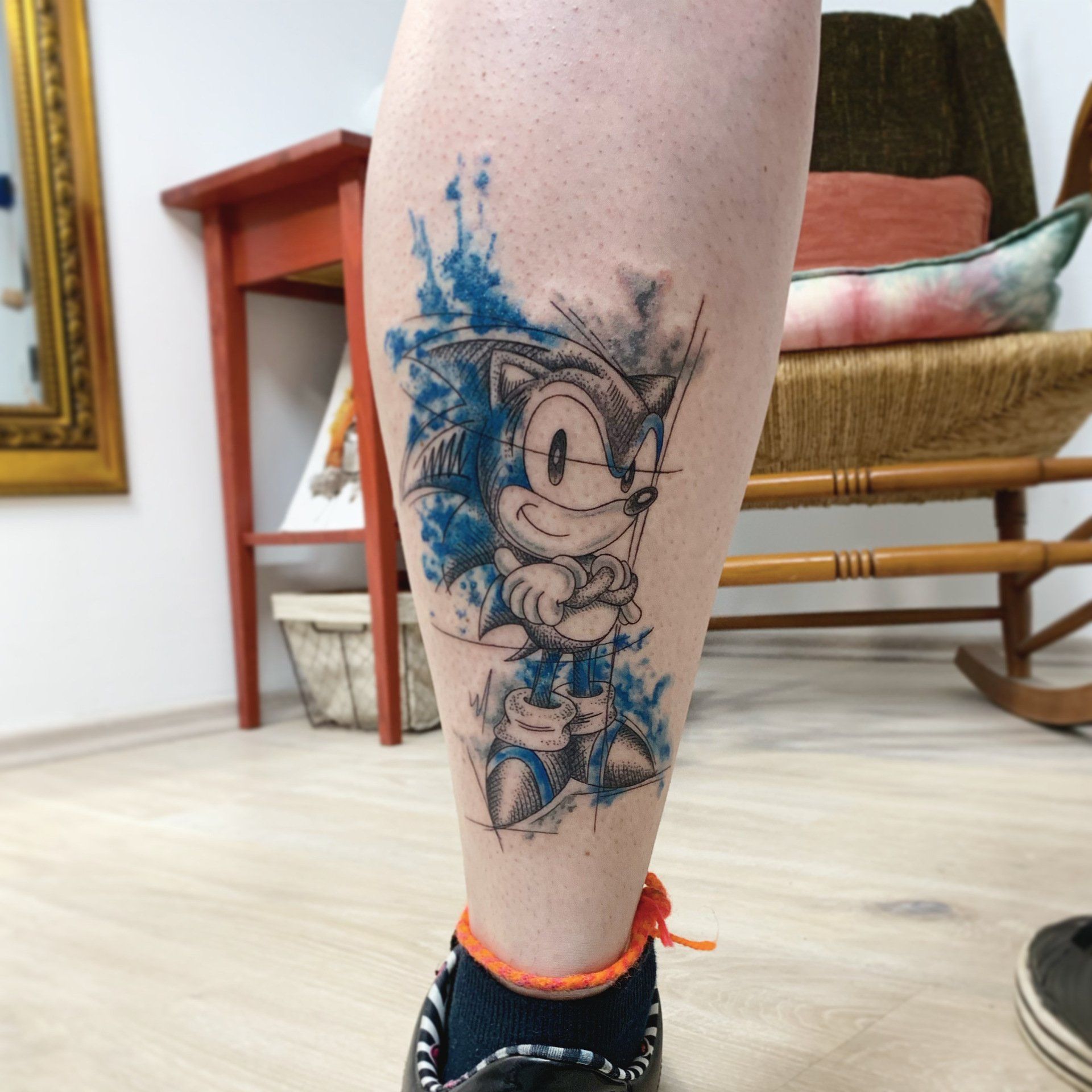 Tattoo von Sonic the Hedgehog auf einem Bein, blauer Hintergrund im Aquarellstil.