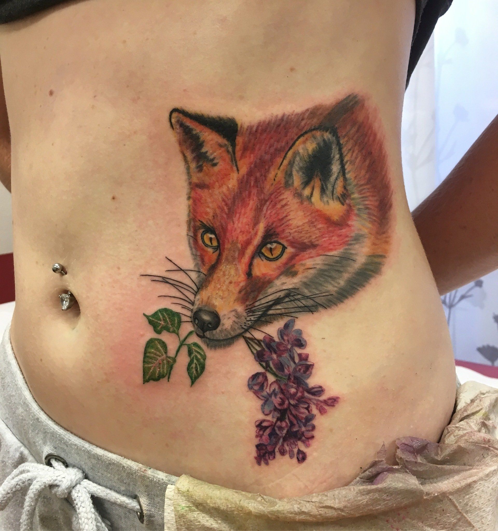 Tätowierung eines Fuchses, der lila Blumen im Maul hält, auf dem Bauch einer Person.