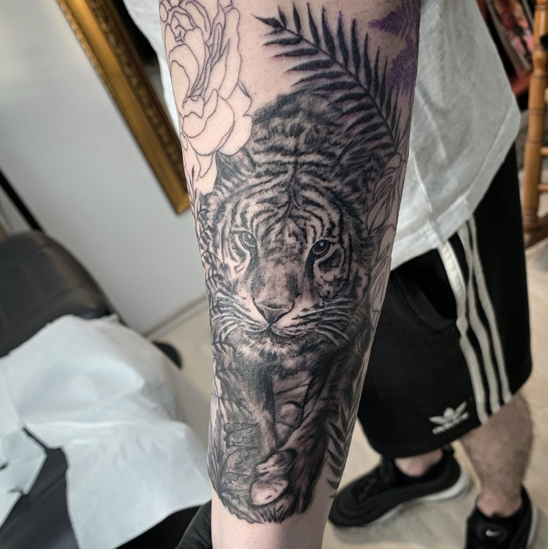 Tätowierung eines Tigers mit detailliertem Gesicht auf einem Arm, umgeben von Blättern und Blumen. Schwarz-weiß.