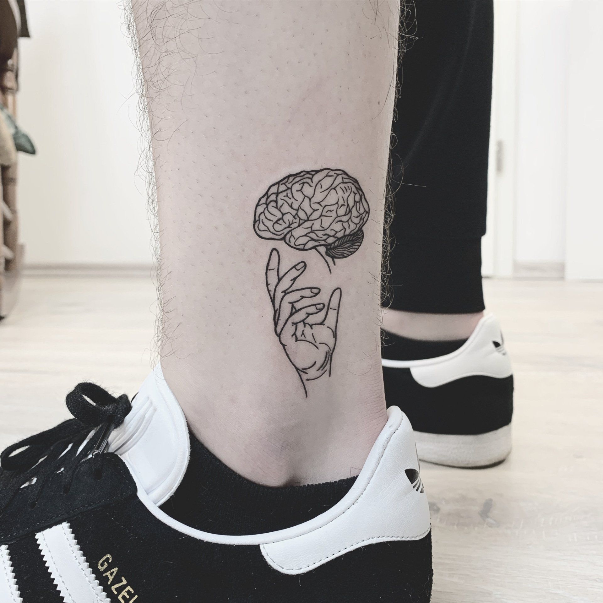 Knöcheltattoo: Hand hält ein Gehirn. Person in schwarzen Turnschuhen und Hose in einem Zimmer.