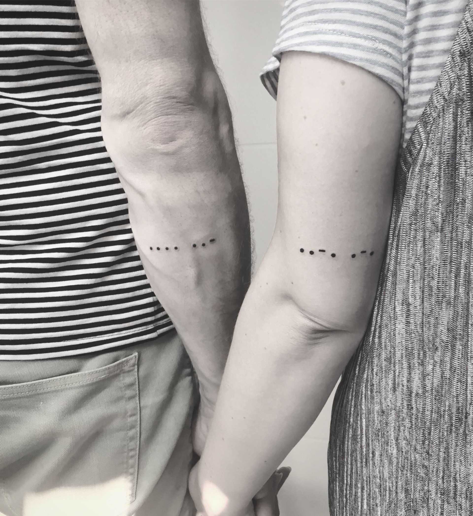 Zwei Arme halten Händchen, jeder mit einem Morsecode-Tattoo. Ein Arm trägt ein gestreiftes Hemd, der andere ein schlichtes Oberteil.