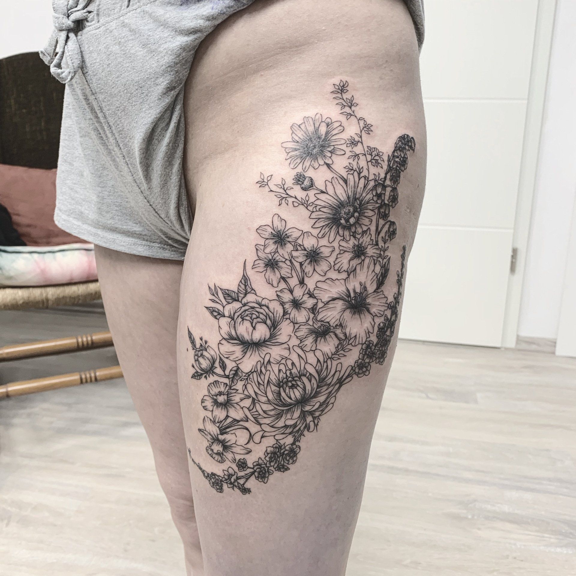 Schwarzes Blumen-Tattoo auf dem Oberschenkel einer Person, sichtbar durch die Shorts. Innenaufnahme, natürliches Licht.