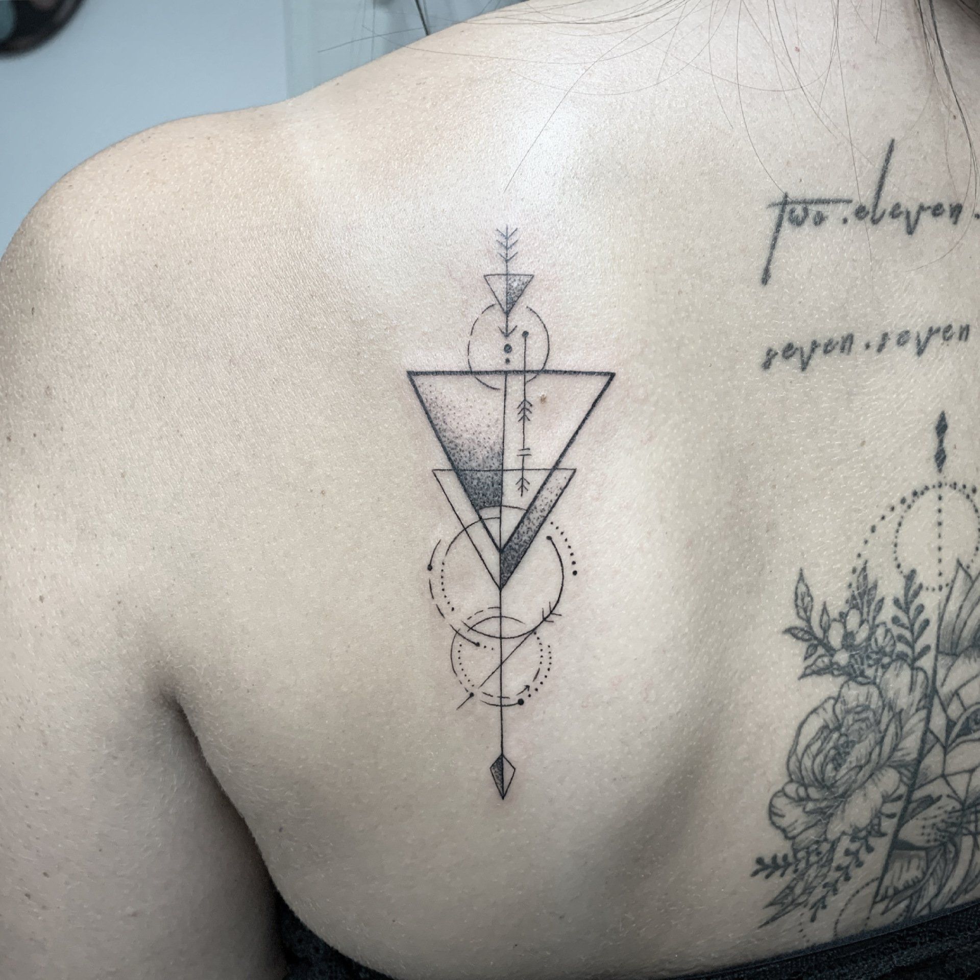 Geometrisches Tattoo auf dem Rücken, bestehend aus Dreiecken, Kreisen und einem kleinen Pfeil, mit einem teilweisen floralen Tattoo.