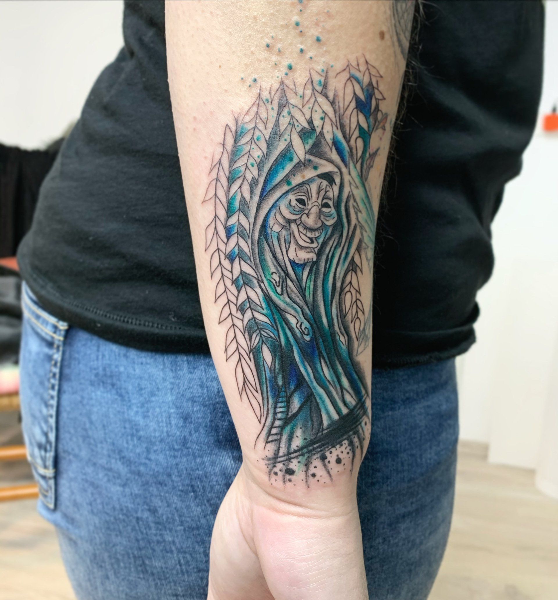 Das Tattoo auf dem Arm zeigt eine blau gekleidete Gestalt mit lächelndem Gesicht, umgeben von Bäumen.