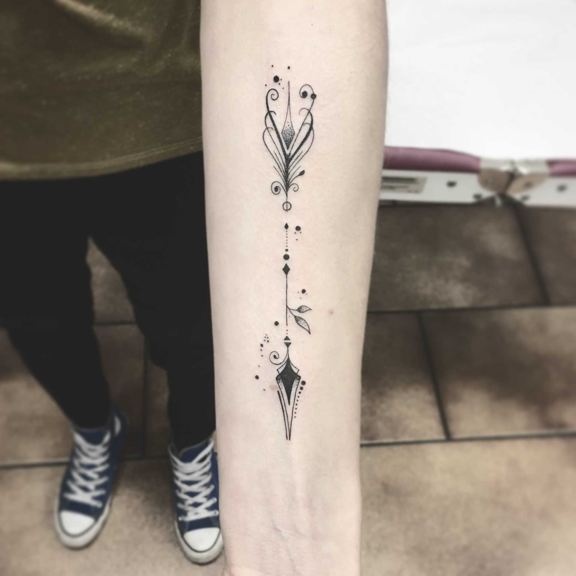 Arm mit schwarzem Tattoo, das ein vertikales Muster aus abstrakten Formen und Punkten zeigt; Person trägt Hose und Turnschuhe.