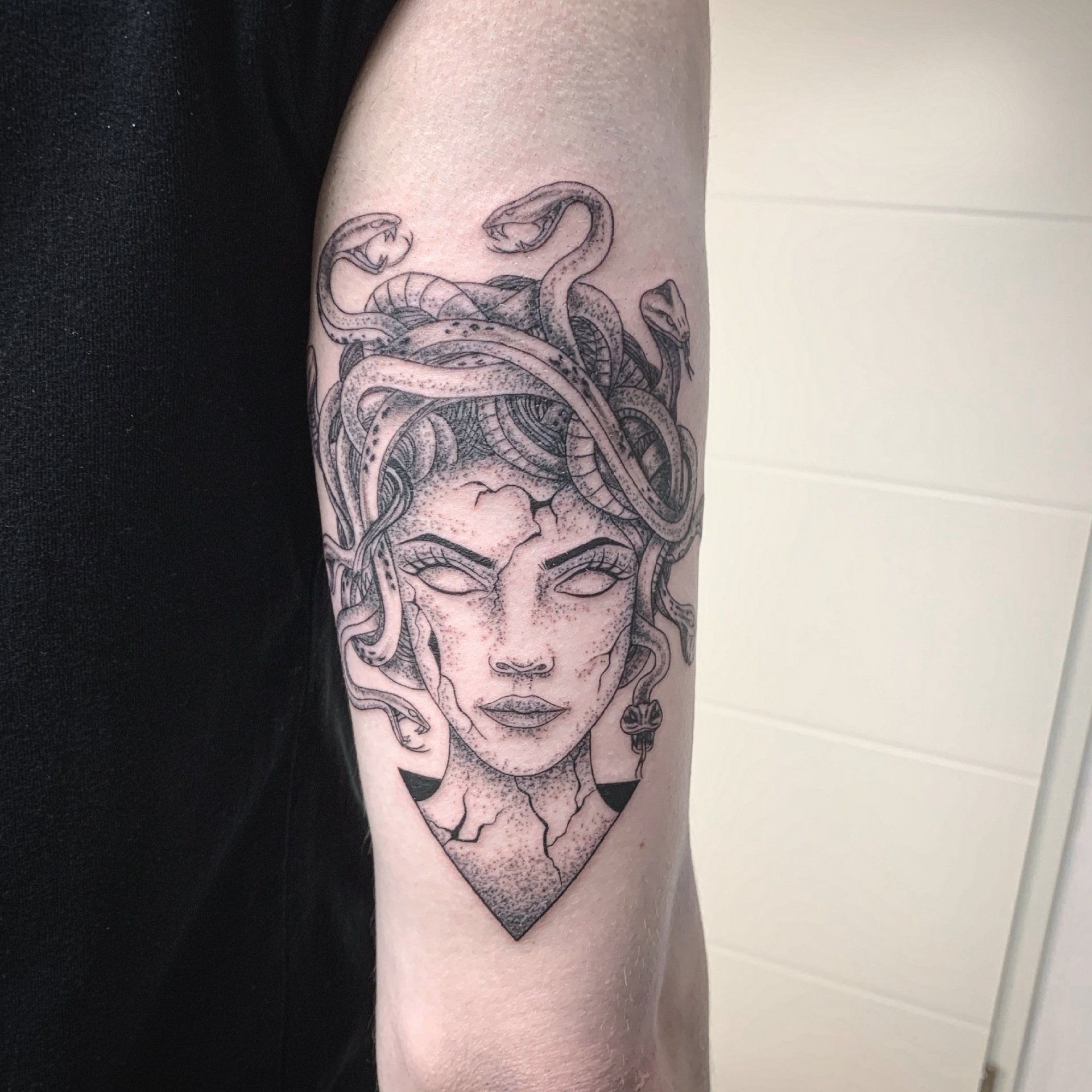 Medusa-Tattoo auf dem Arm einer Person, in Schwarz und Grau dargestellt, mit Schlangen als Haare und einem dreieckigen Sockel.