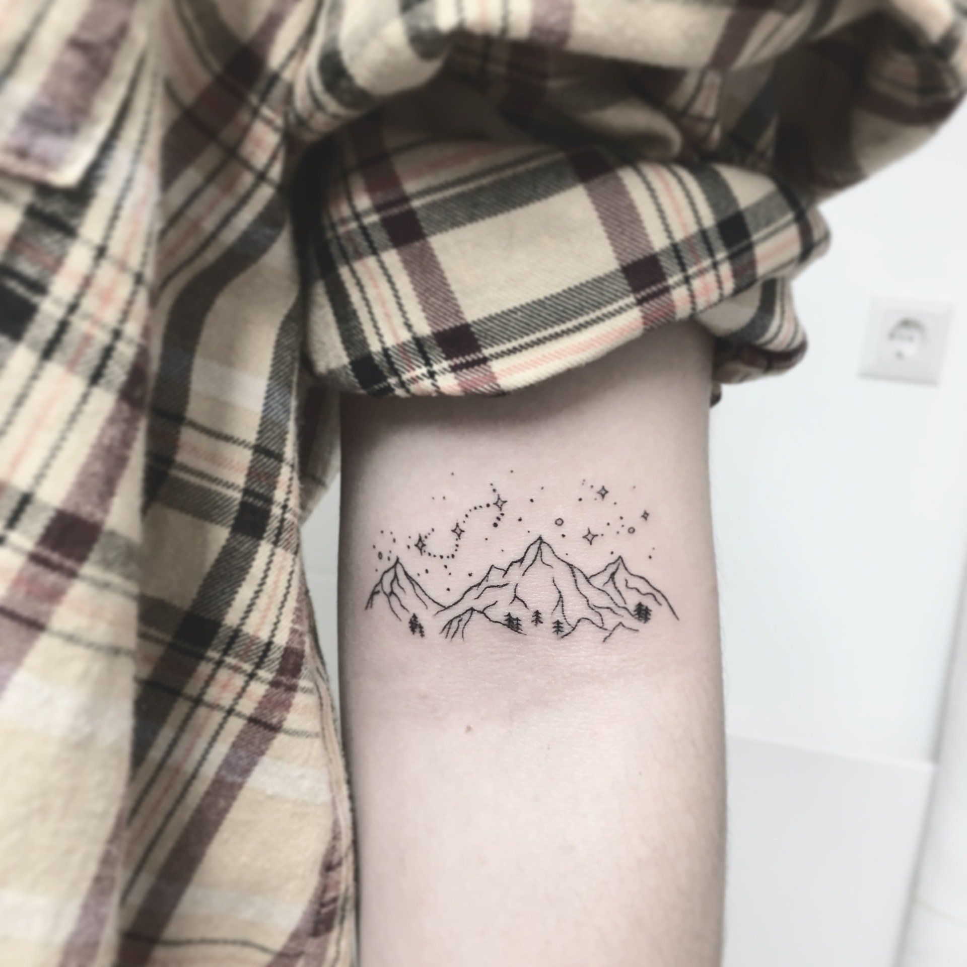 Tätowierung einer Bergkette mit Sternen auf dem Arm, Person trägt ein kariertes Hemd.