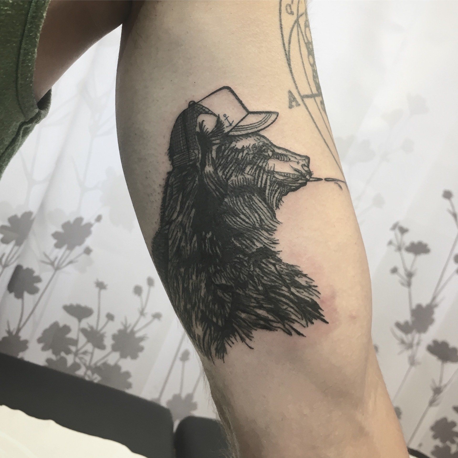 Tätowierung auf dem Arm eines Bären mit Hut; schwarze Schattierung, im Profil gezeichnet.