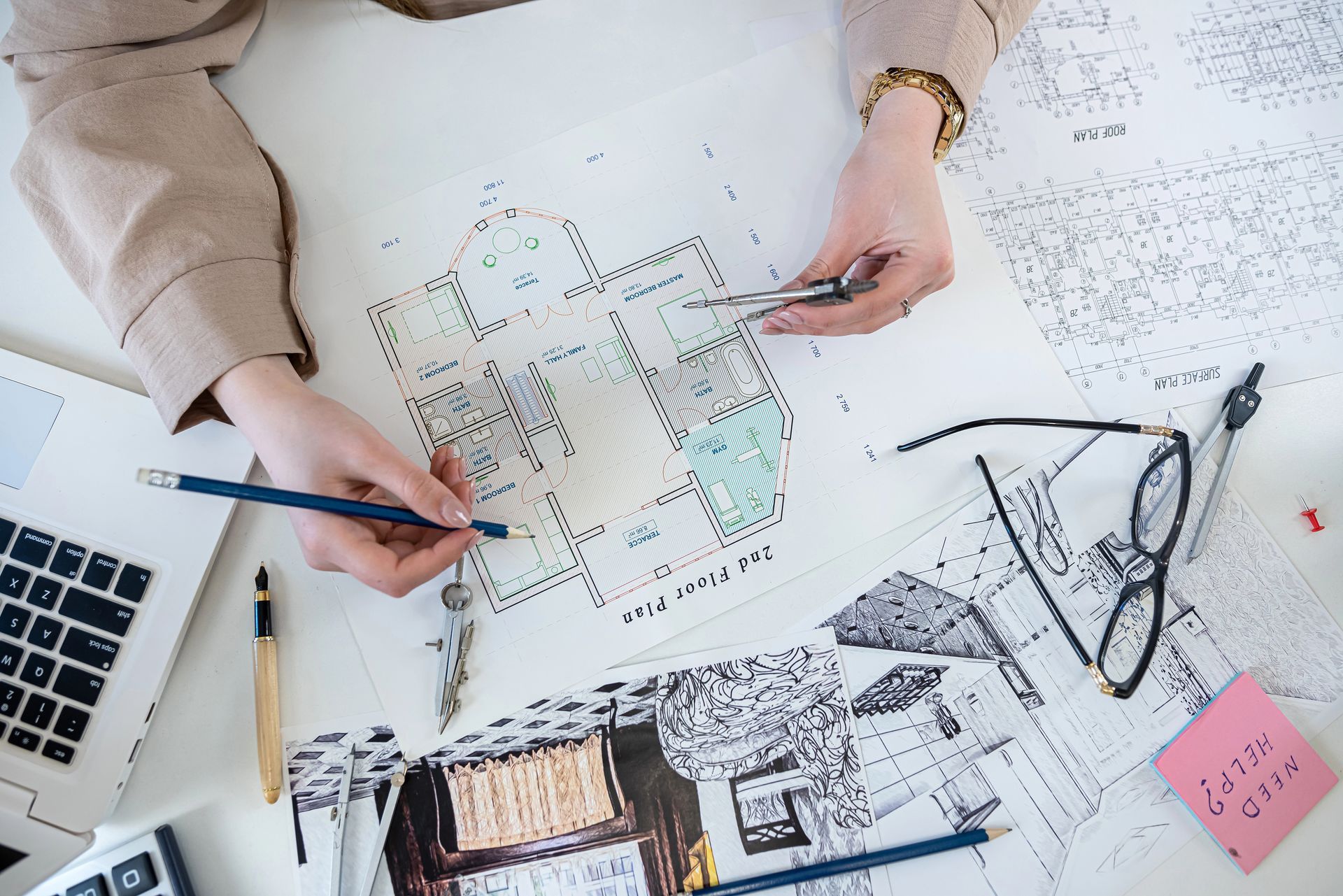Mani che abbozzano progetti architettonici su una scrivania con computer portatile, righello, occhiali e disegni sparsi.