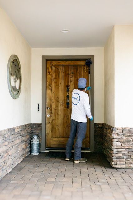 Person in white shirt and blue cap knocking on a wooden front door under a covered entryway