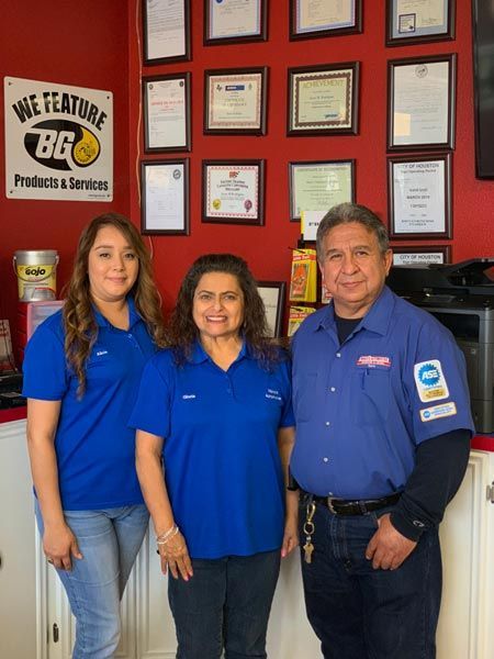 Three employees in blue shirts posing in front of framed certificates and a company sign. | Mario’s Automotive