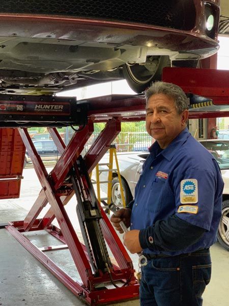 Mechanic in blue shirt stands under a lifted red car in an auto repair shop. | Mario’s Automotive