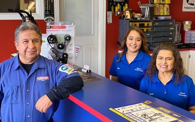 Three staff in blue uniforms stand behind a reception counter in an auto repair shop. | Mario’s Automotive