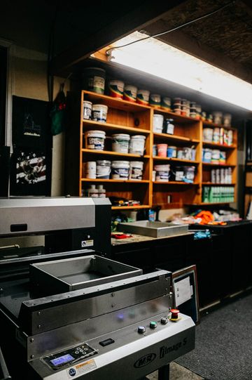 A cluttered workshop with paint cans on shelves, a printing machine in front.