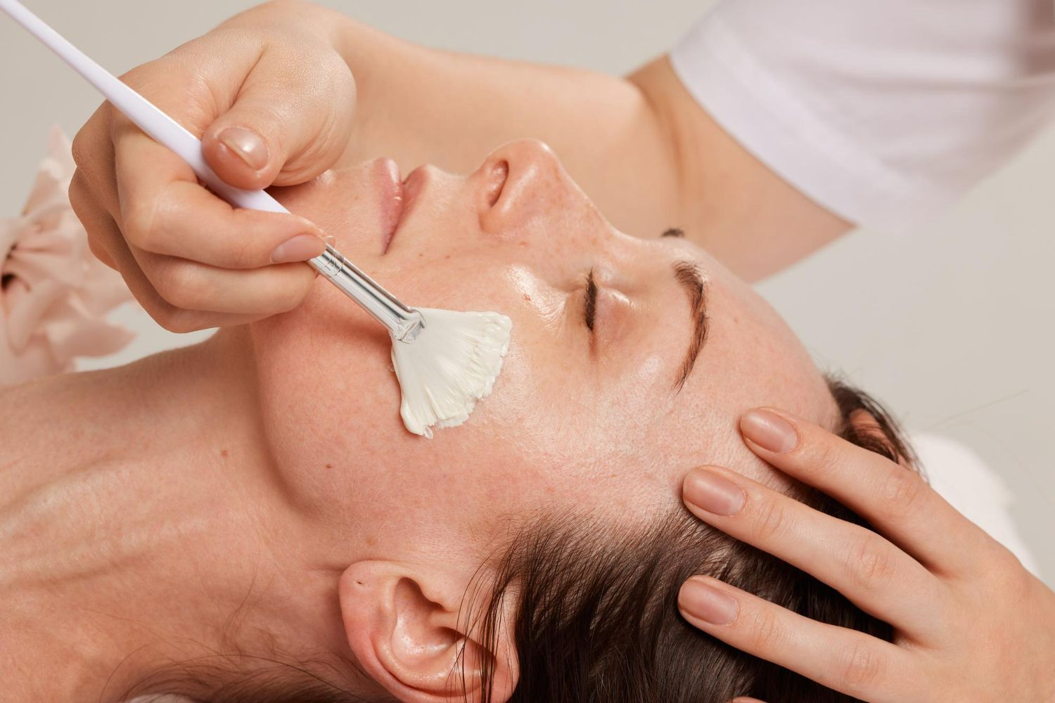 A person receives a facial treatment, with a technician applying a white cream to their cheek using a fan brush.