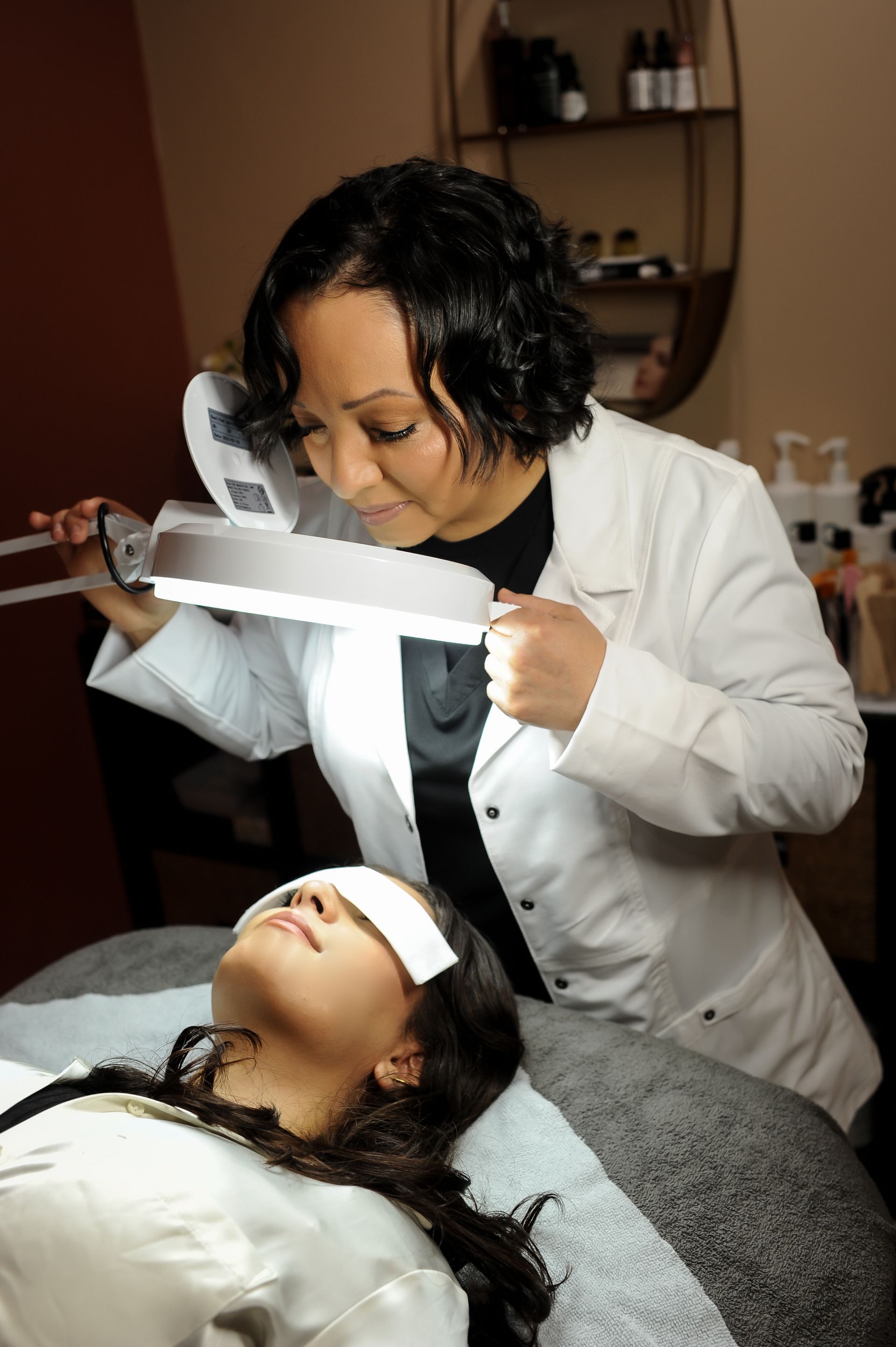 A technician wearing a white coat uses an illuminated magnifying lamp to examine a client lying on a spa treatment table.