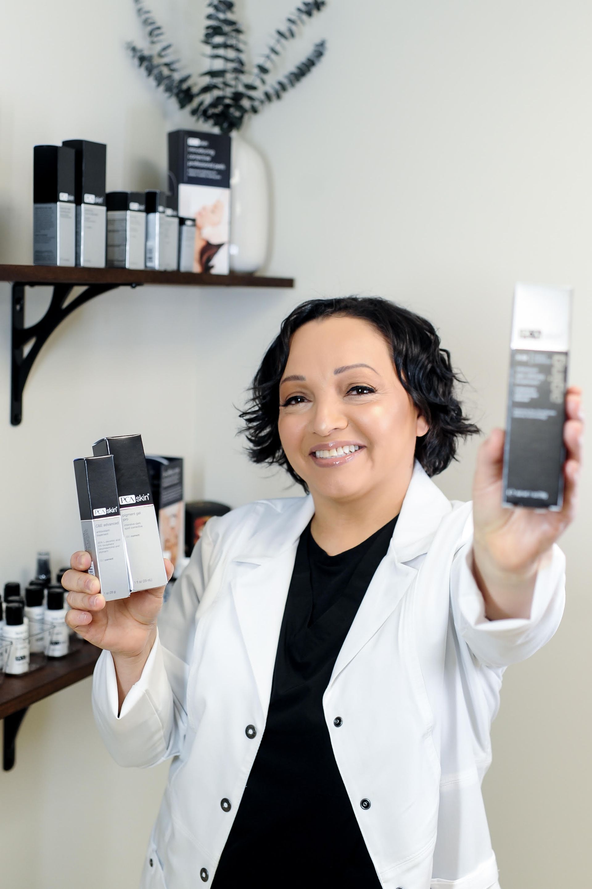 A professional in a white coat smiling and holding skincare products in front of a shelf displaying various beauty items.