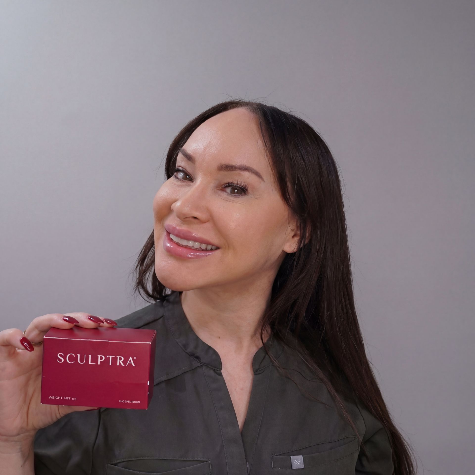 A person with dark hair smiling while holding a small, red box labeled